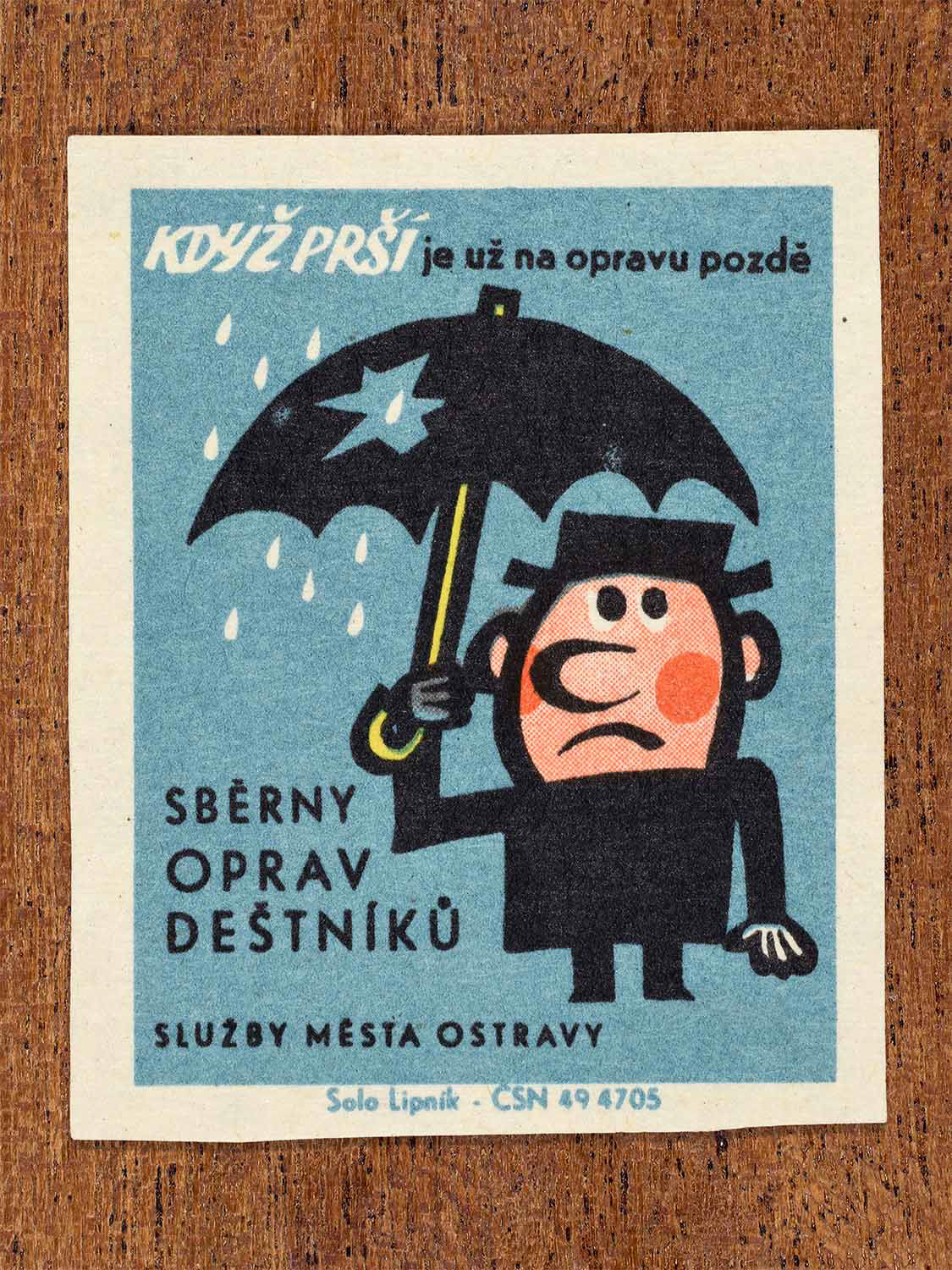 Vintage 1967 Czech matchbox label showing cartoon a man under a leaking umbrella in the rain, on a blue background. Produced by the Solo Lipník match factory to promote municipal umbrella repair services.