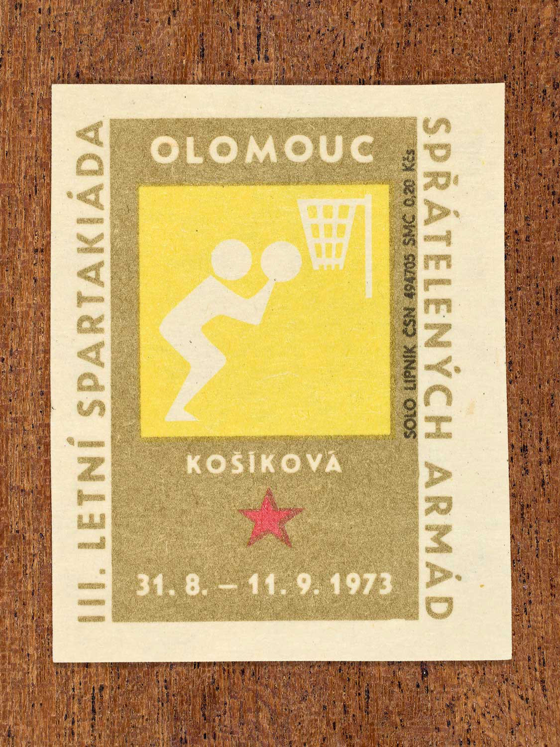 Vintage 1973 Czech matchbox label showing a simplified white basketball player leaping toward a hoop, on a yellow and gold background. Produced by the Solo Lipník match factory to promote physical training and demonstrate collective military readiness among allied armies.