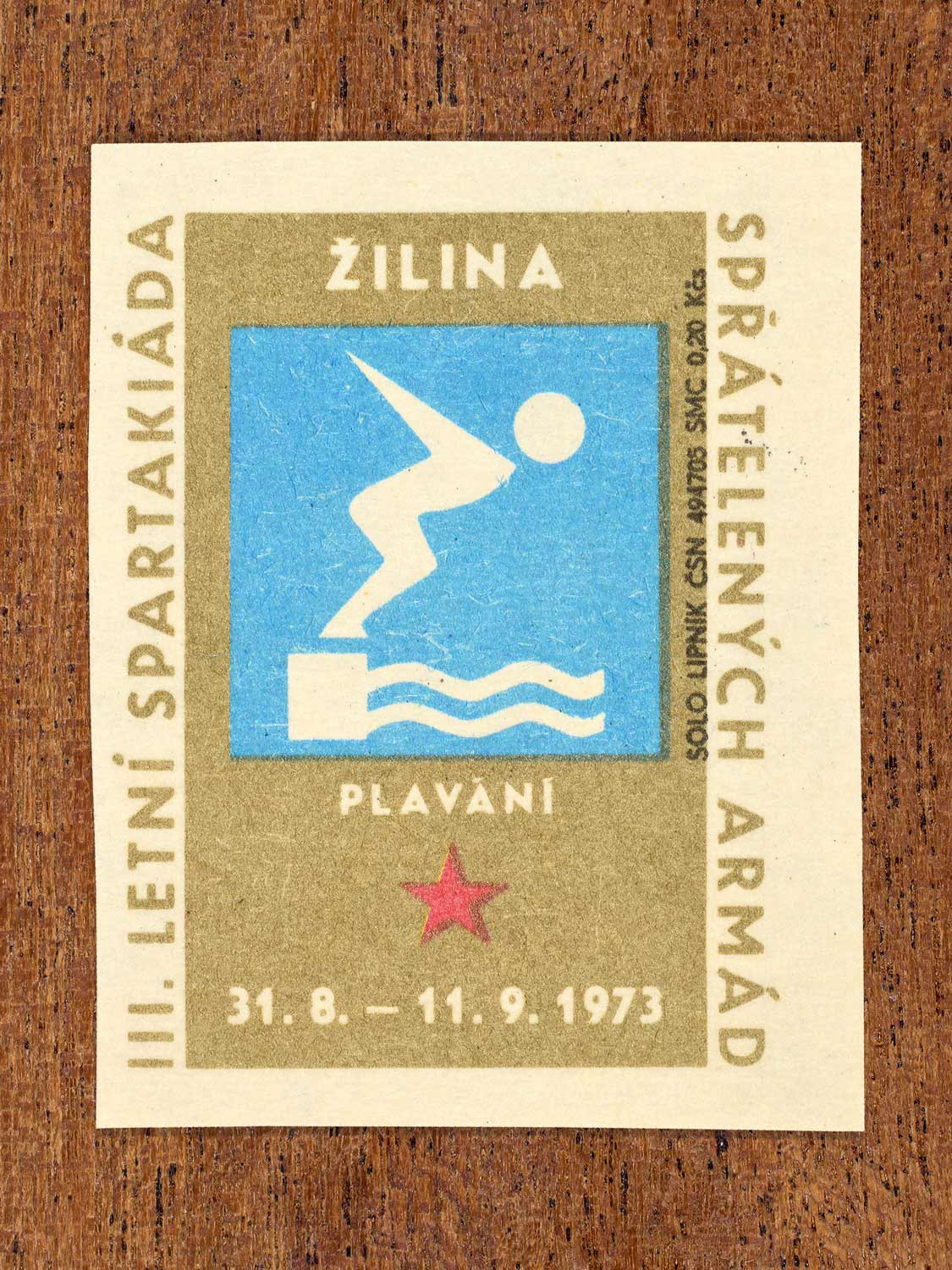 Vintage 1973 Czech matchbox label showing a simplified white swimmer figure on a blue and gold background. Produced by the Solo Lipník match factory to promote physical training and demonstrate collective military readiness among allied armies.