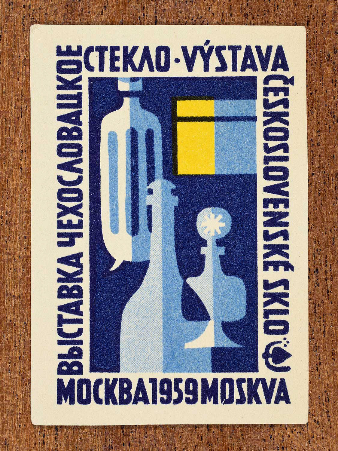 A 1959 Czechoslovak matchbox label featuring a mid-century modern illustration of glass vessels and bottles against a deep blue background. The central graphic is bordered by bilingual Russian and Czech text promoting a glass exhibition in Moscow.