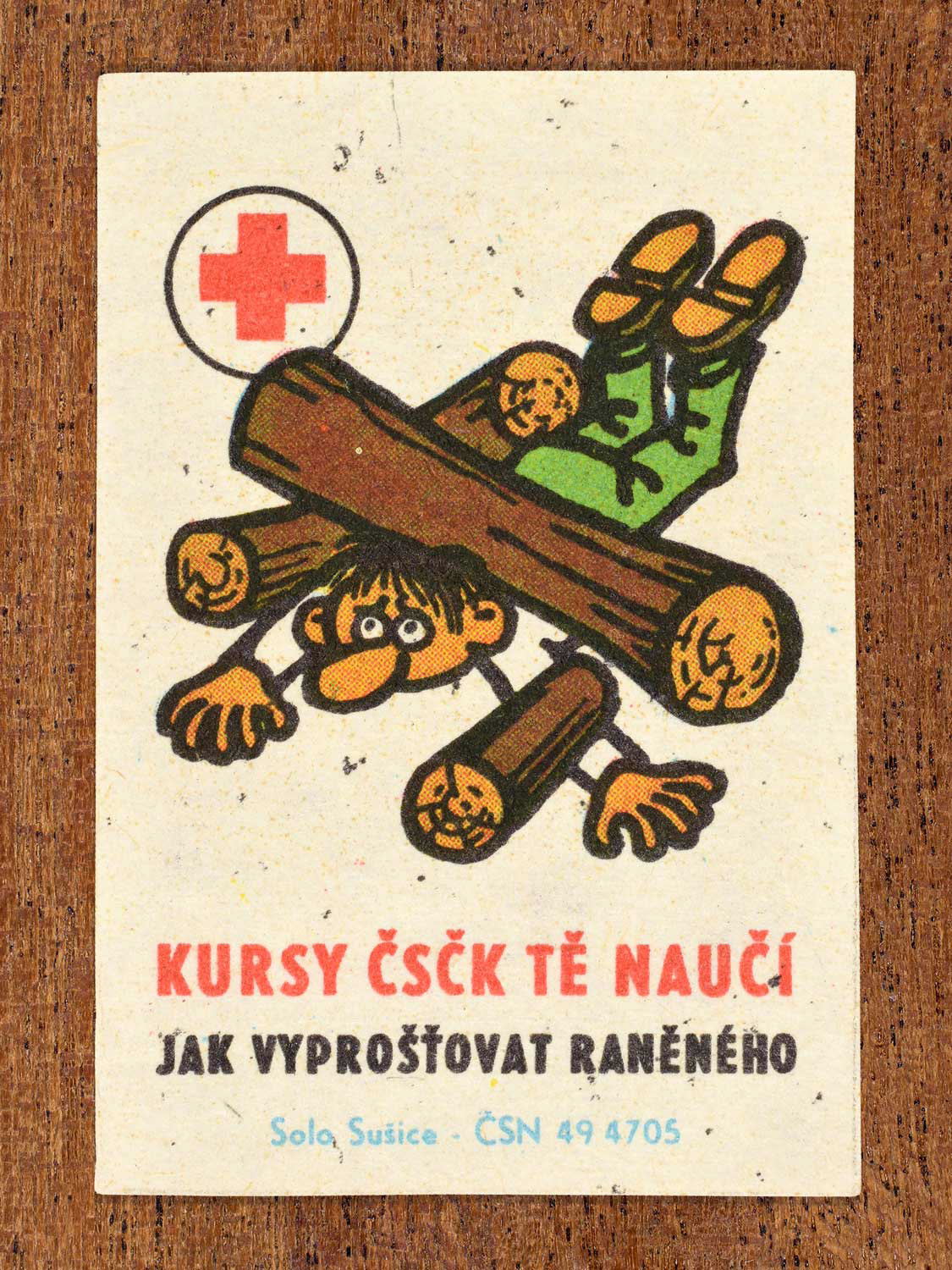 Vintage c. 1966 Czech matchbox label showing an injured person trapped beneath heavy logs. Produced by the Solo Sušice match factory to promote first-aid education and safe extrication of injured persons in cooperation with the Czechoslovak Red Cross.