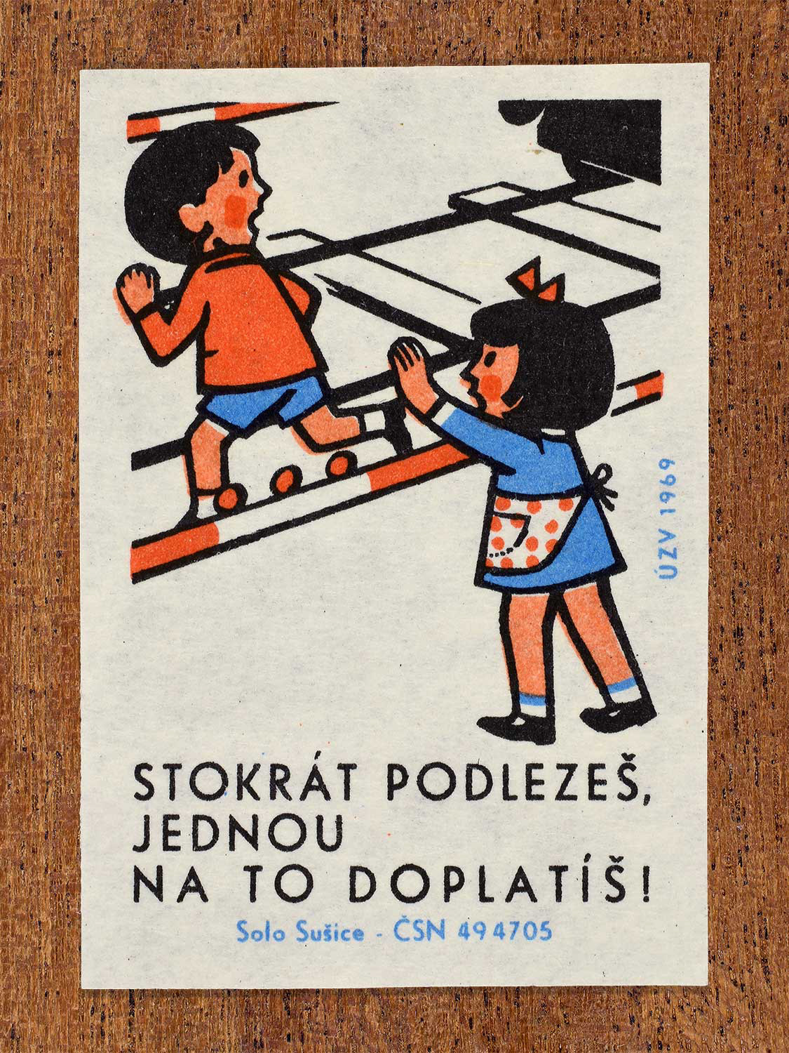 Vintage 1969 Czech matchbox label showing a young boy crawling under a lowered railway crossing barrier while a young girl watches. Produced by the Solo Sušice match factory to promote safe and responsible behavior around railway crossings.