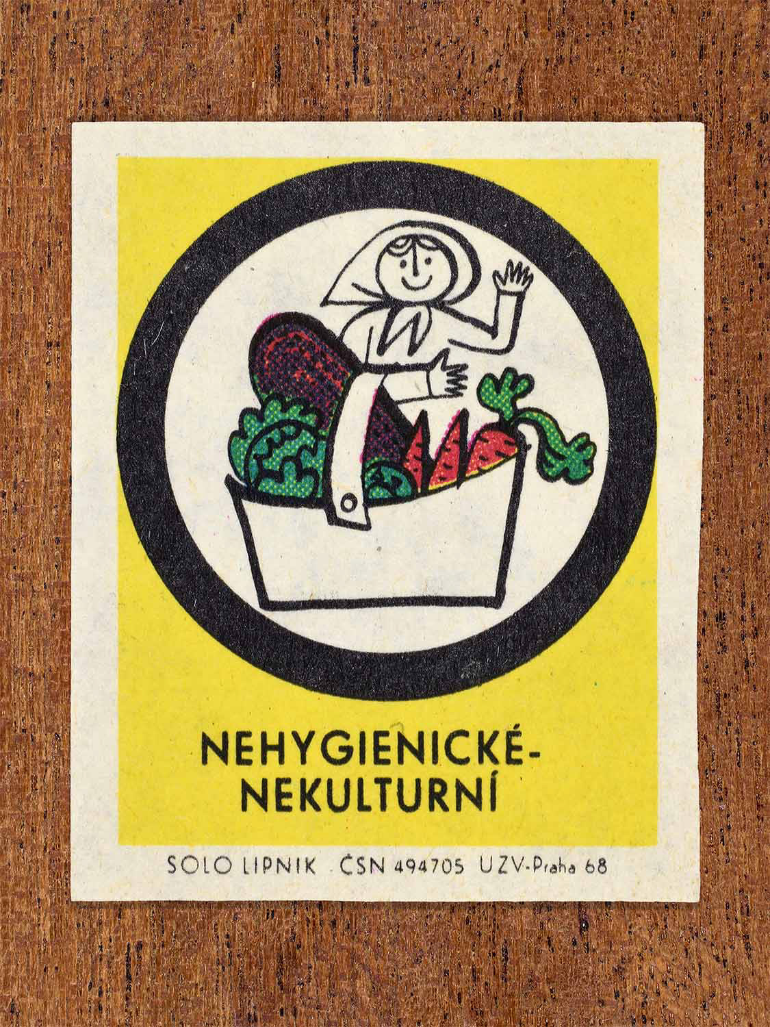 Vintage c. 1968 Czech matchbox label showing a person holding a basket of vegetables, framed in a circle and on a yellow background. Produced by the Solo Lipník match factory to criticise unhygienic handling of food.