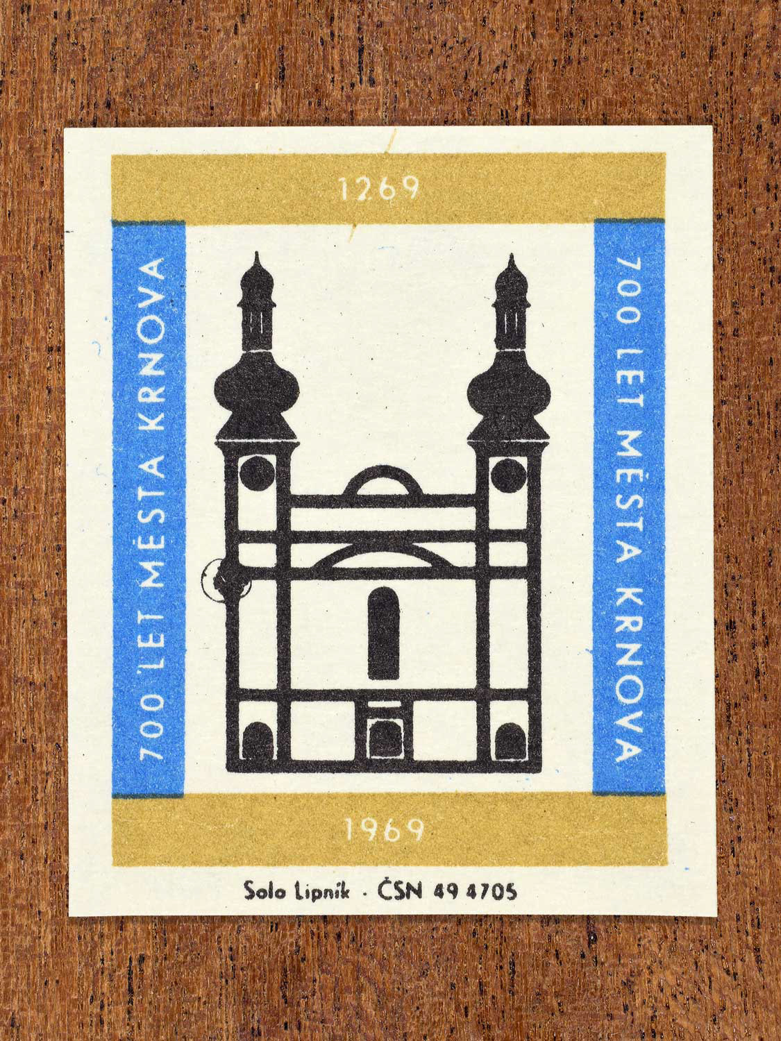 Vintage 1969 Czech matchbox label showing the Church of the Nativity of the Virgin Mary in Krnov in black with blue and gold framing. Produced by the Solo Lipník match factory to commemorate 700 years of the city of Krnov.