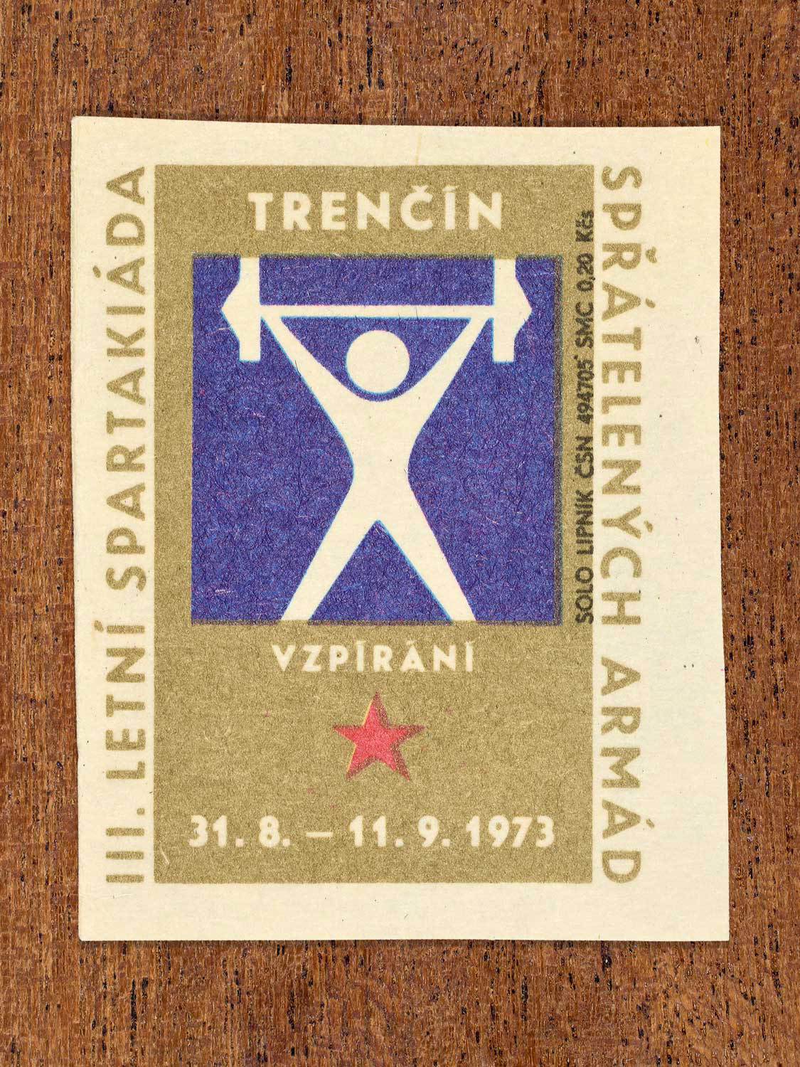 Vintage 1973 Czech matchbox label showing a simplified white figure lifting a barbell above their head, on a blue and gold background. Produced by the Solo Lipník match factory to promote physical training and demonstrate collective military readiness among allied armies.