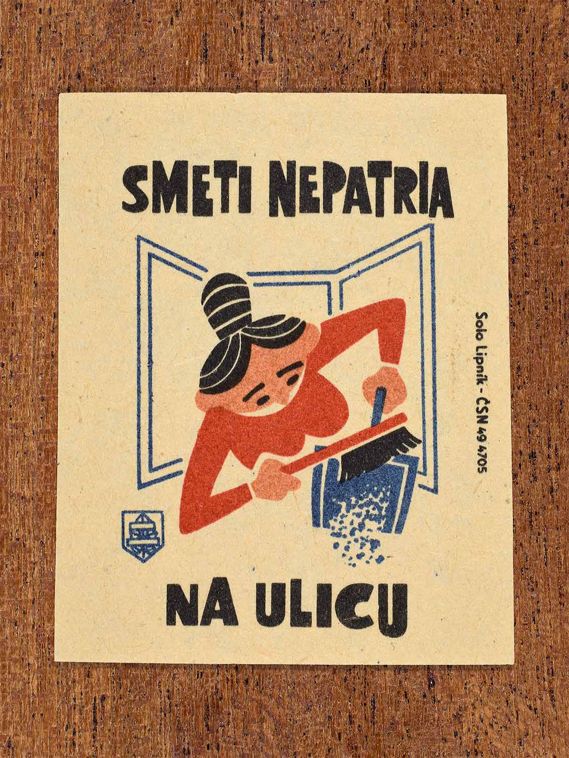 Vintage 1964 Czech matchbox label showing a woman sweeping rubbish out of a window, in blue, red and black colours. Produced by the Solo Lipník match factory to discourage throwing waste onto public streets.