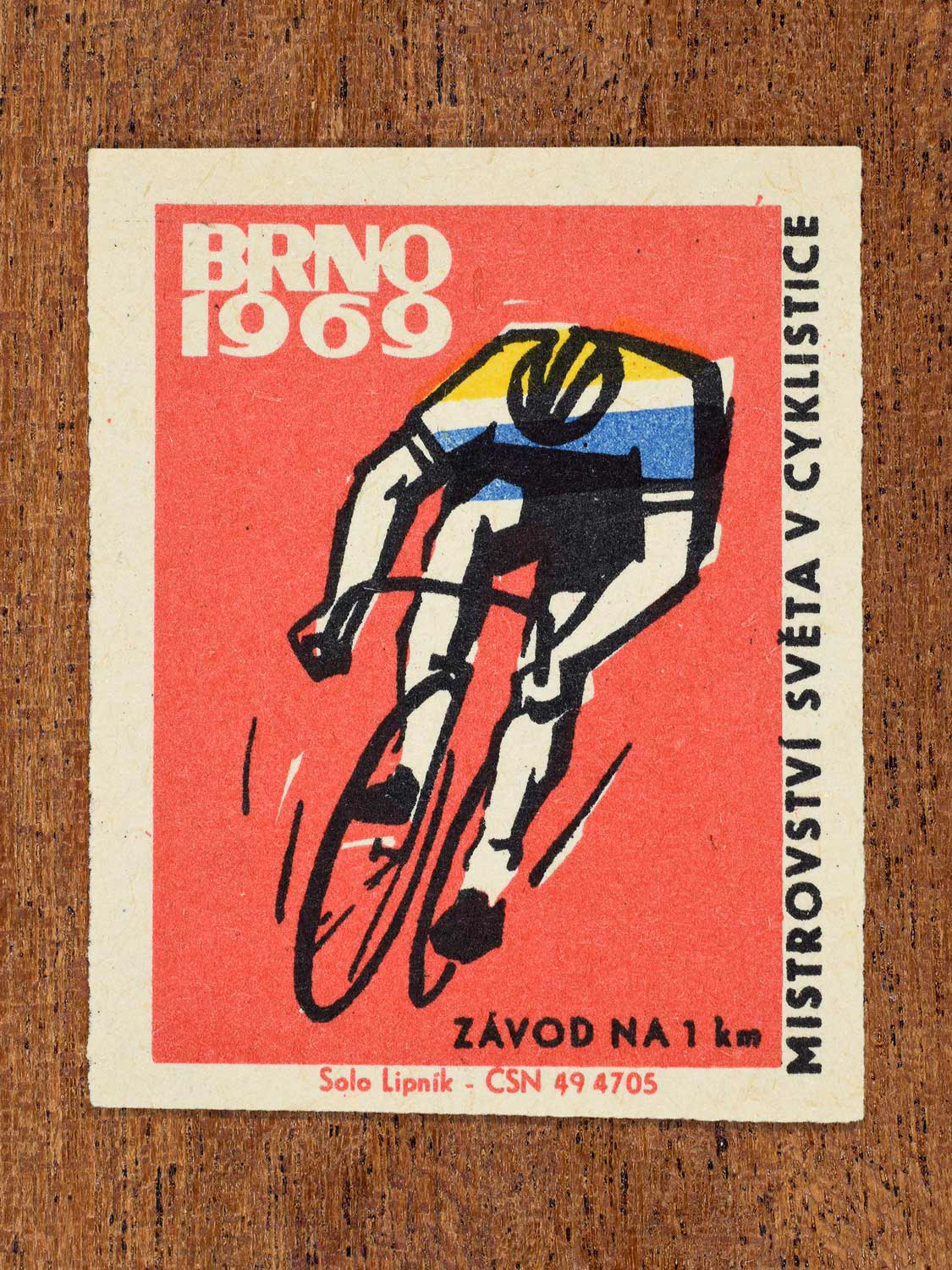 Vintage 1969 Czech matchbox label showing a stylised cyclist racing in a 1 km event, on a red background. Produced by the Solo Lipník match factory to promote cycling at a championship in Brno, Czechoslovakia.