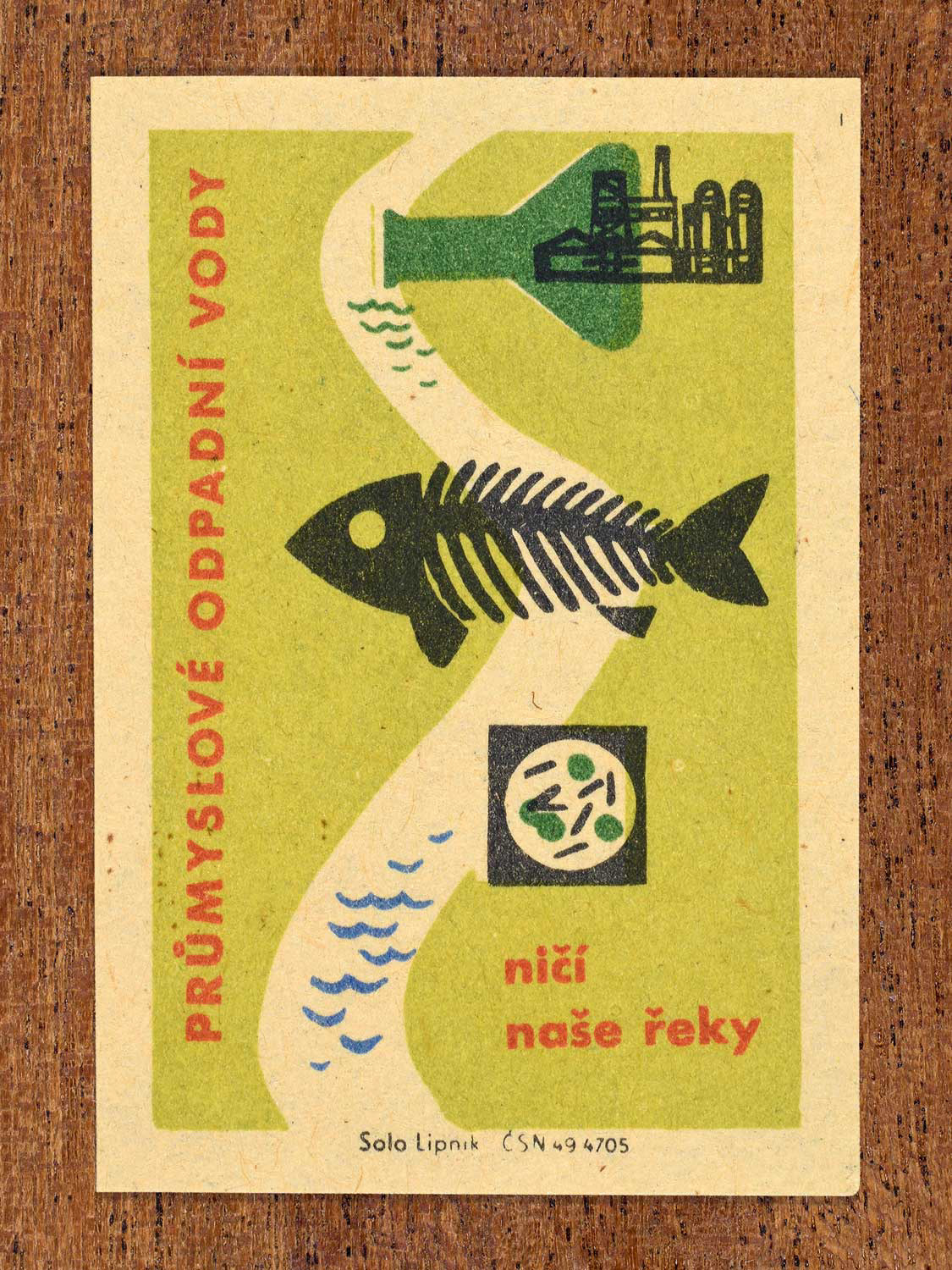 Vintage c. 1963 Czech matchbox label showing a fish skeleton in a polluted river next to factories, in green, red and black colours. Produced by Solo Lipník match factory warning that industrial wastewater destroys rivers.