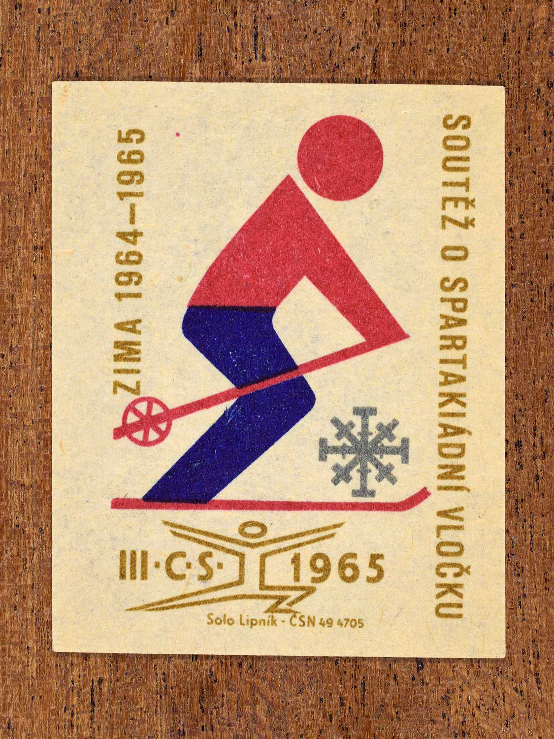 Vintage 1965 Czechoslovak matchbox label showing a winter athlete in motion. Produced by the Solo Lipník match factory to promote Spartakiad athletic performance.