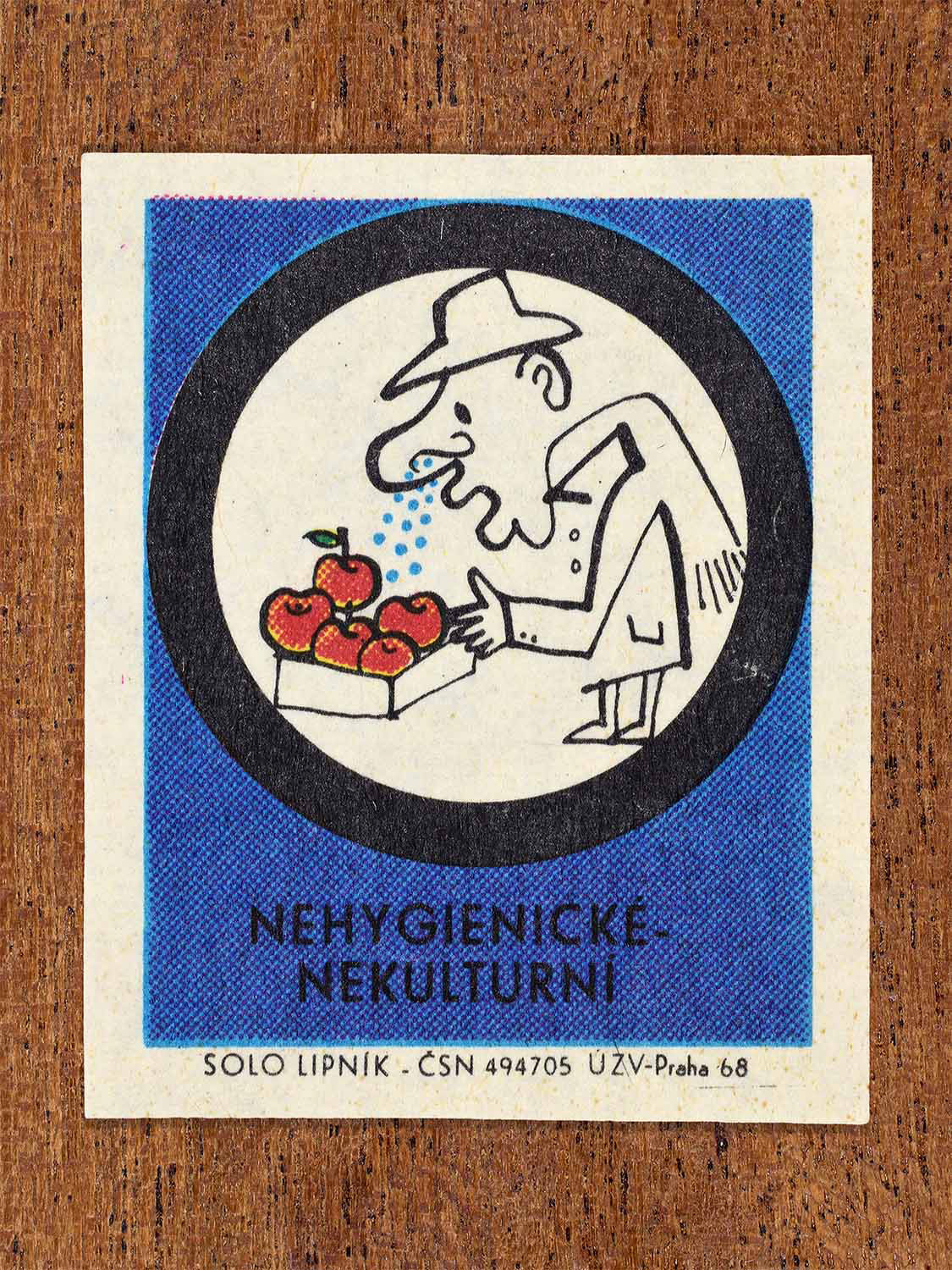 Vintage c. 1968 Czech matchbox label showing a man sneezing or coughing over uncovered fruit, framed in a circle and on a blue background. Produced by the Solo Lipník match factory to warn against unhygienic food contamination.