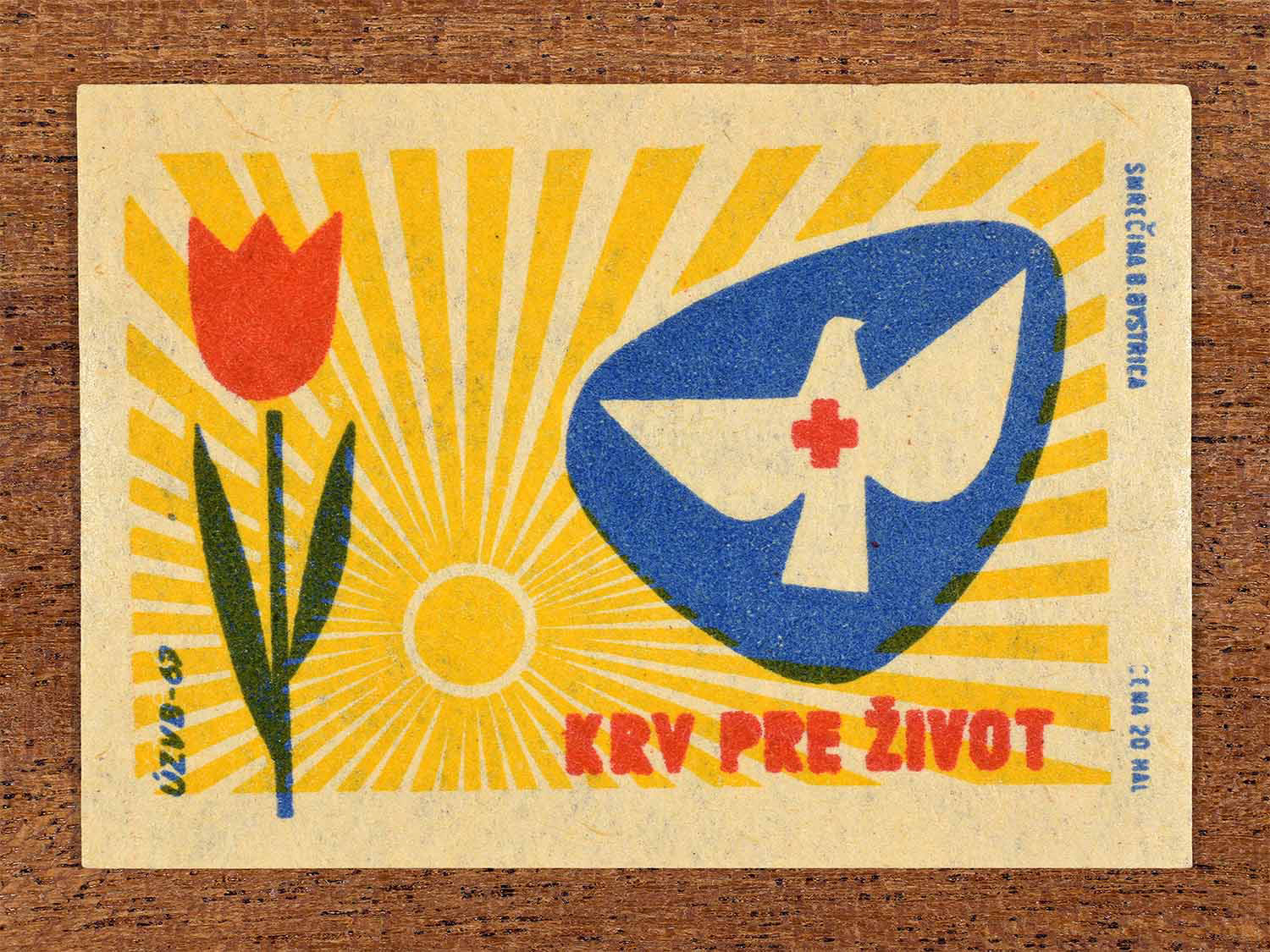 Vintage 1967 Czech matchbox label showing a red tulip flower with sun rays behind it, and a white dove health symbol. In red, blue and yellow colours. Produced by the Smrečná match factory, Banská Bystrica, to promote blood donation.