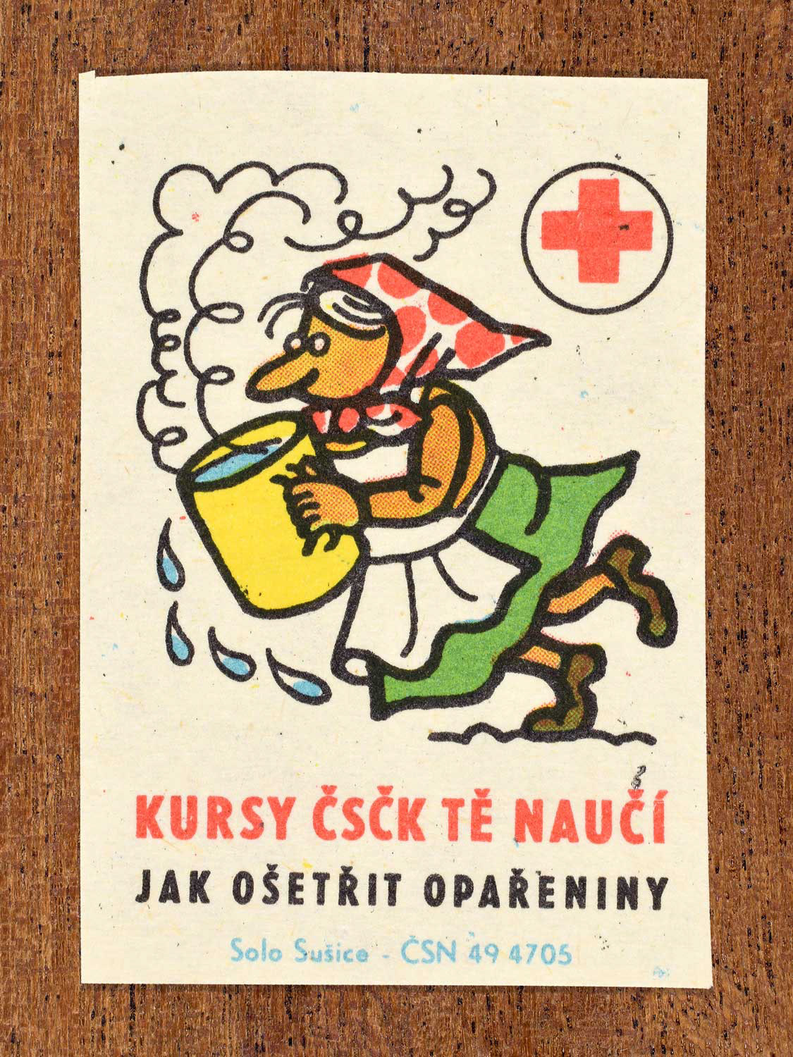 Vintage c. 1966 Czech matchbox label showing a woman tripping while holding a large pan of boiling water. Produced by the Solo Sušice match factory to promote first-aid education and proper treatment of scald injuries in cooperation with the Czechoslovak Red Cross.