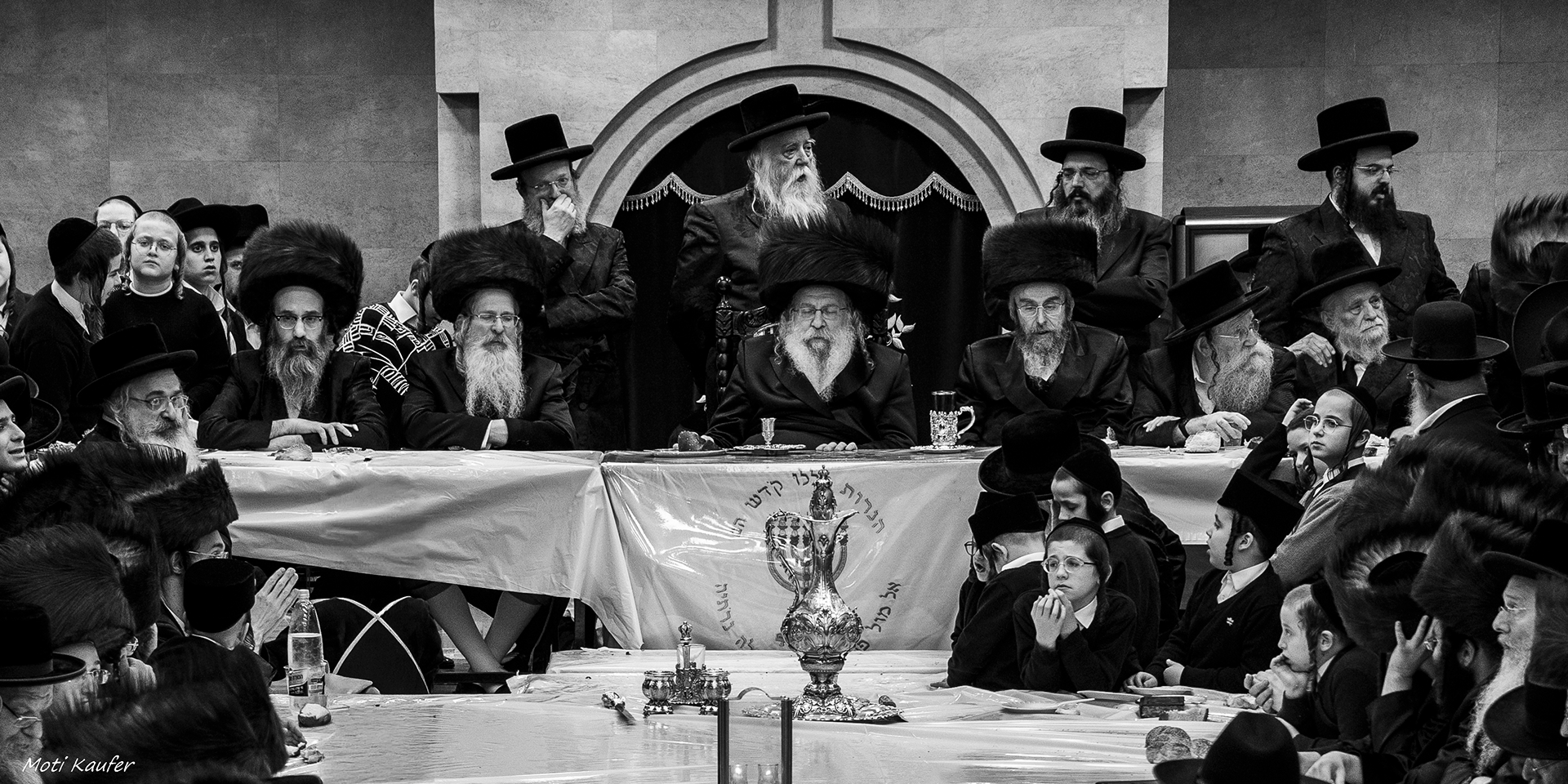 Hasidic Jews gather around their Rabbi’s table for a symbolic meal, where they listen attentively to the Rabbi’s Torah teachings. The gathering is also marked by communal singing, creating a deeply spiritual and communal atmosphere.