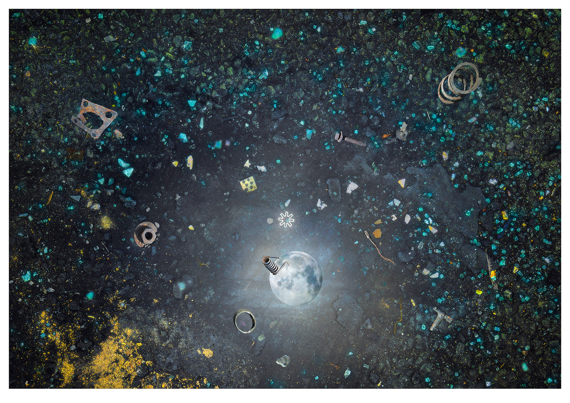 The original photograph is a tarmacadam road with lots of broken windscreen glass. To this  metal pieces have been added as floating debris orbiting around a distant blue planet/moon. The illusion resembles floating debris in space.