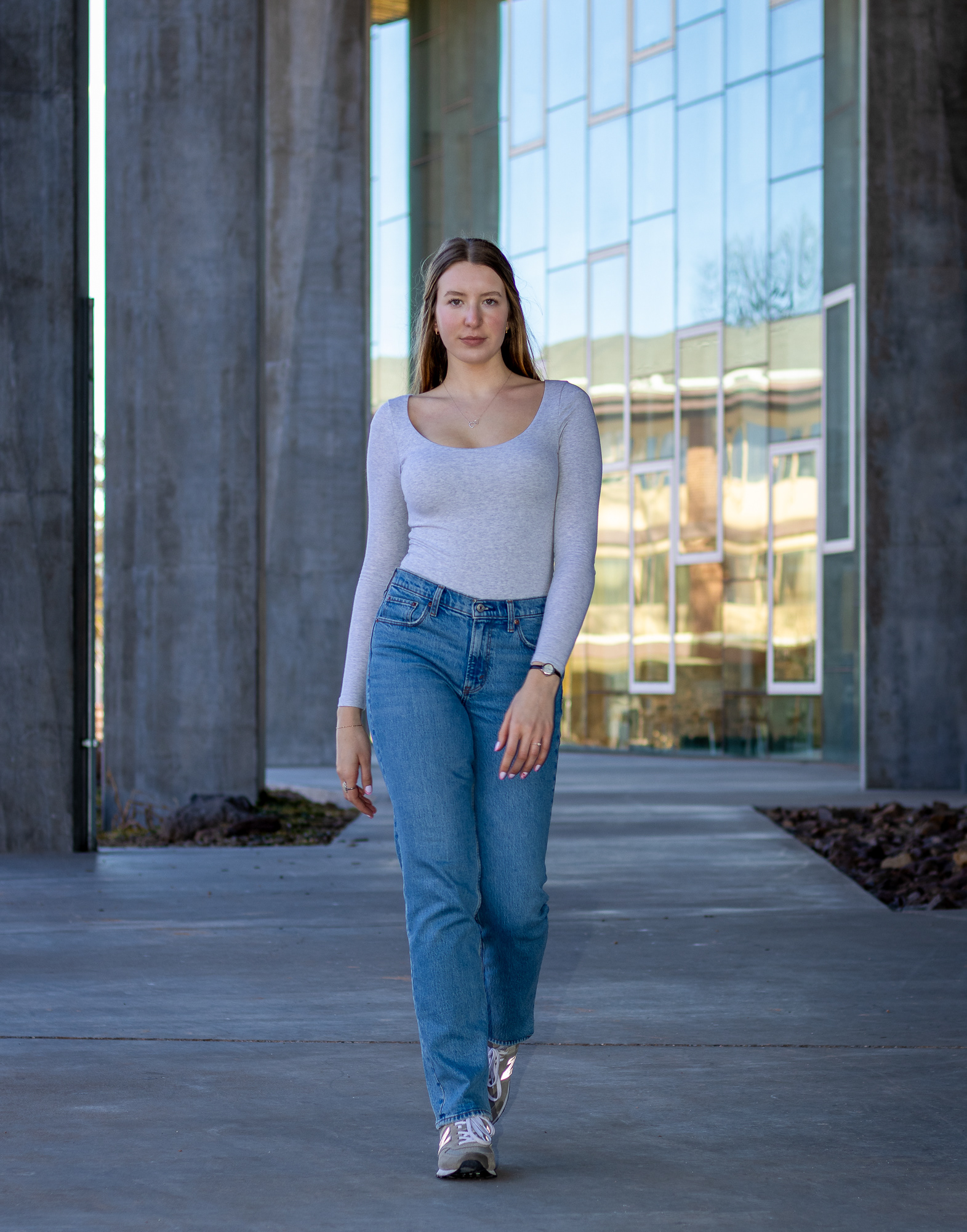 Woman in jeans and grey long sleeve top walking confidently past a glass building