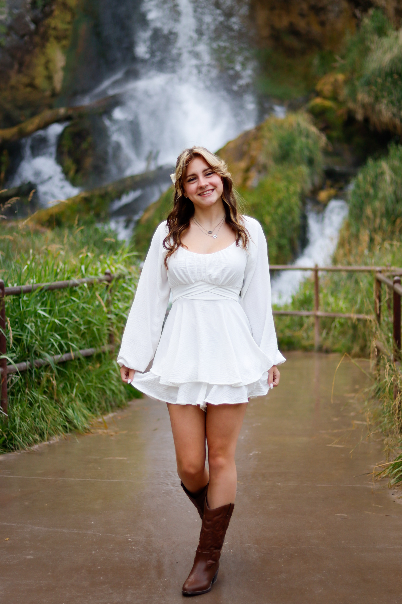 Woman in white dress and boots walking on a trail in front of a waterfall