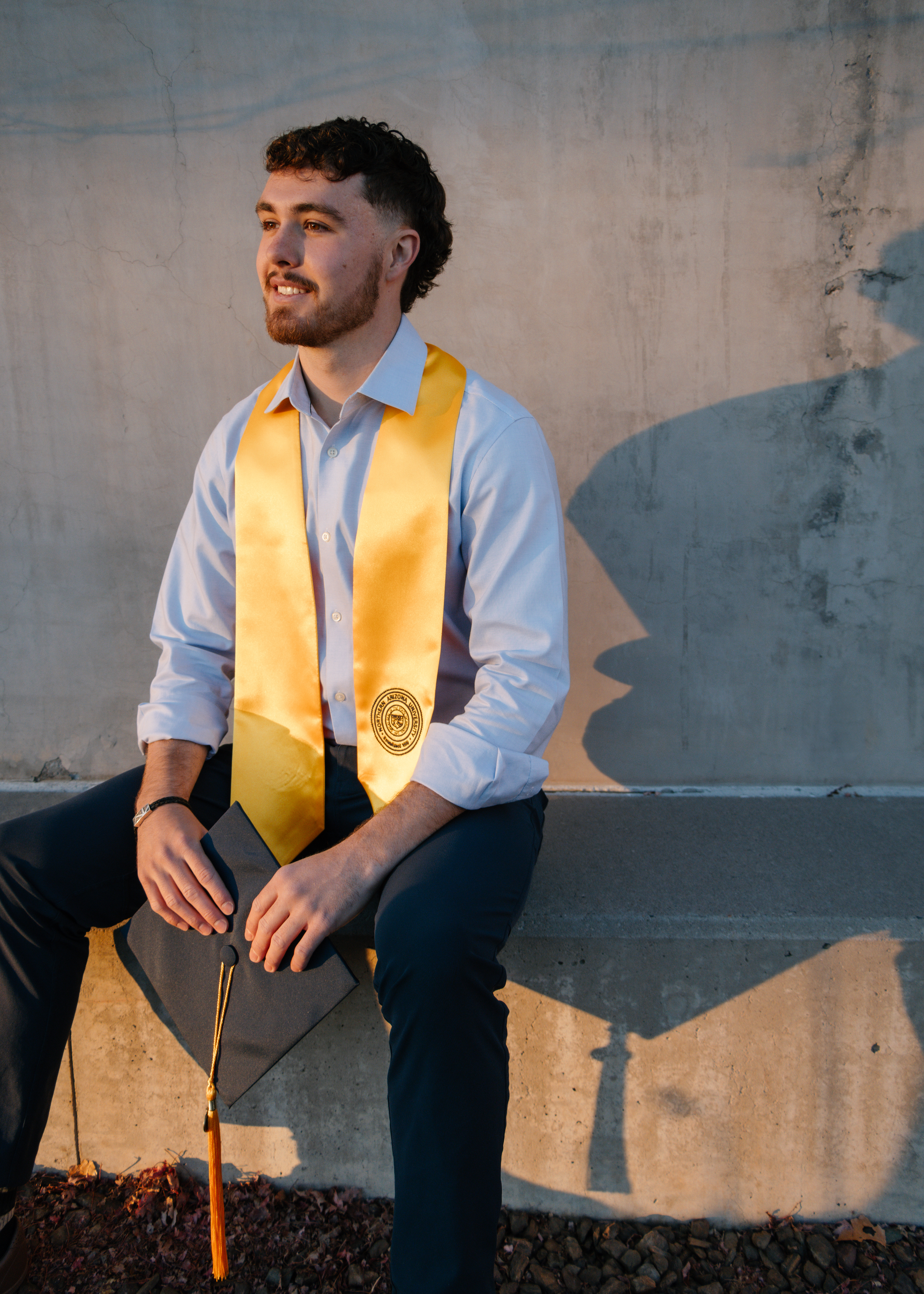 Man in graduation attire sitting on a concrete step, highlighted by an afternoon sun with his shadow behind him