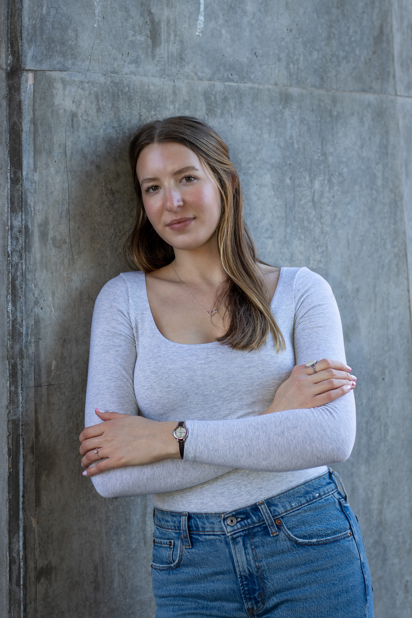 Woman in long sleeve grey shirt leaning casually with arms crossed against a concrete wall