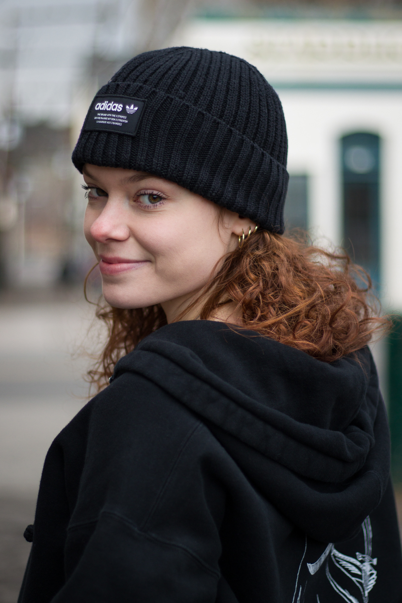 Woman in a hoodie and beanie smiling over her shoulder, urban setting