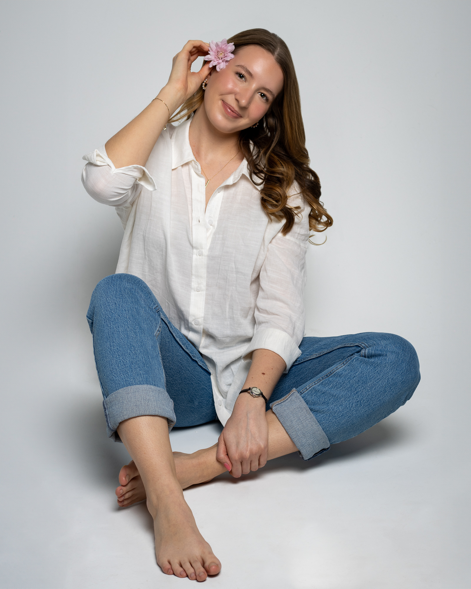 Woman sitting barefoot in jeans and a white shirt against a pale backdrop holding a flower in her hair