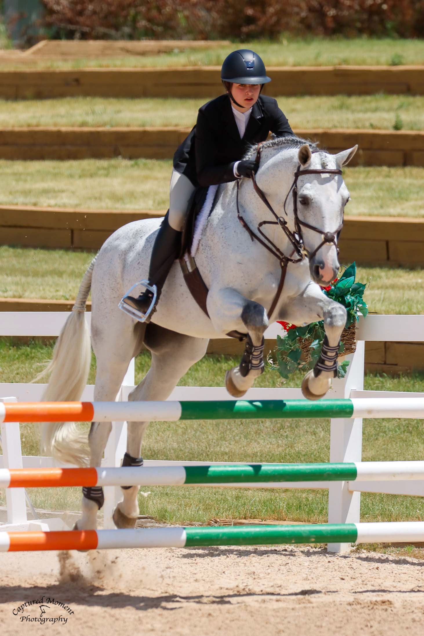 Rider guiding a grey horse over a green and orange vertical jump
