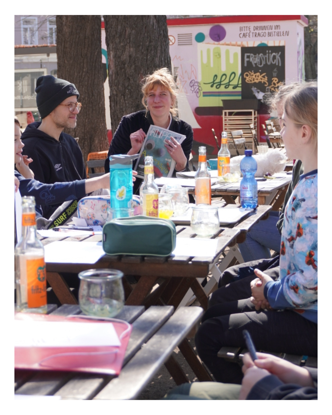 Die Kinderredaktion der Kinderzeitung Flippo beim Workshop im Garten – mit Wiebke Steinert, die die Ausgabe „Anderssein“ in den Händen hält, und Friedemann Lichtenthal. Auf dem Tisch stehen Limonadeflaschen, Stifte und Notizzettel liegen verstreut. Im Hintergrund: das Café Trago.