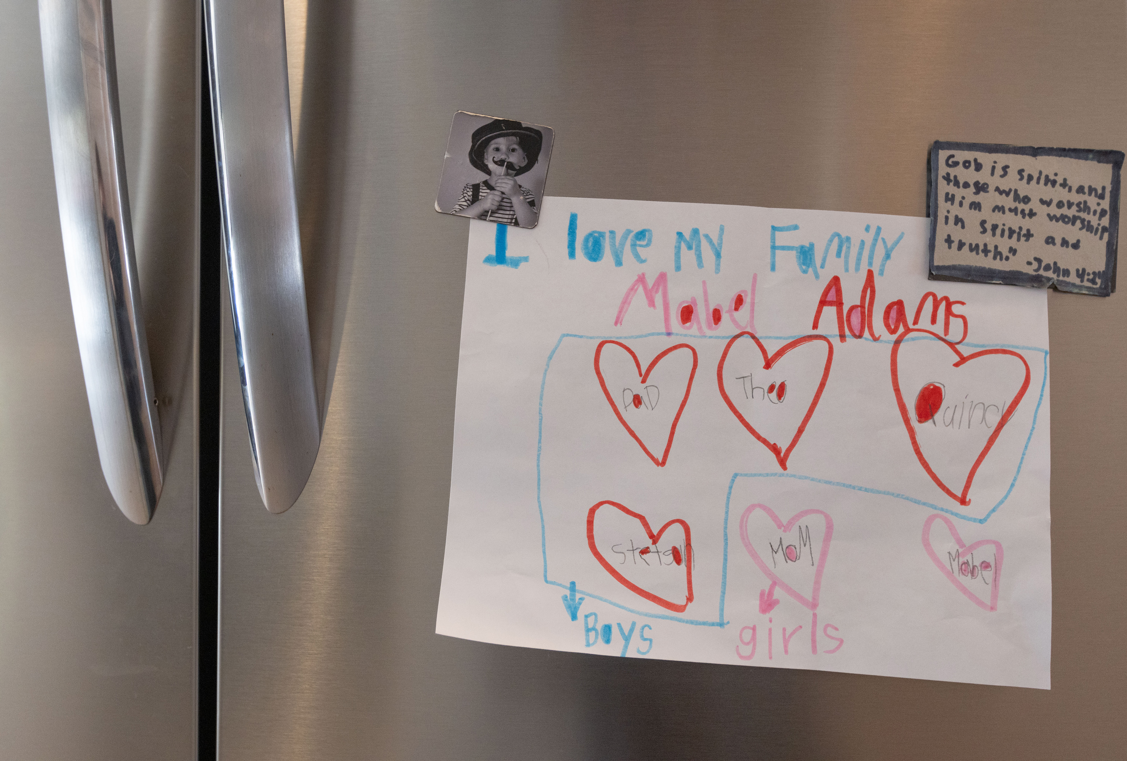 A piece of artwork by Mabel Adams (6) reading "I love my family" is hung on the fridge at the Adams house in Monroe, Ga on Feb. 22, 2025. A Bible verse and photo magnet of a young Quincy Adams (13) wearing his father Zach Adams' hat hold up the artwork. 