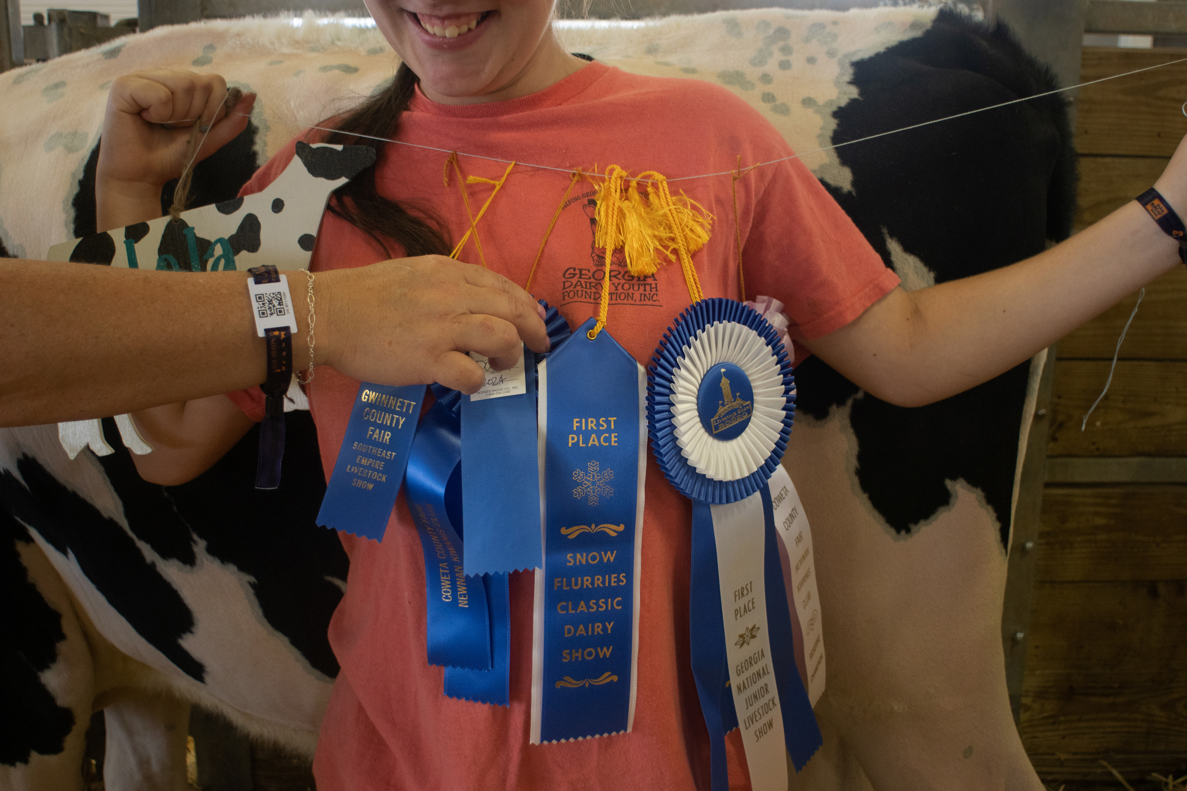 Mia Robinson of Cleveland, Georgia, 16, stands in front of her cow Lola while holding a string of first place ribbons that she's won with her in the past year at the Georgia National Fairgrounds and Agriculture Center on Oct. 5, 2024. Her mother, Heather Robinson, helps her straighten out the ribbons as the wind blows.