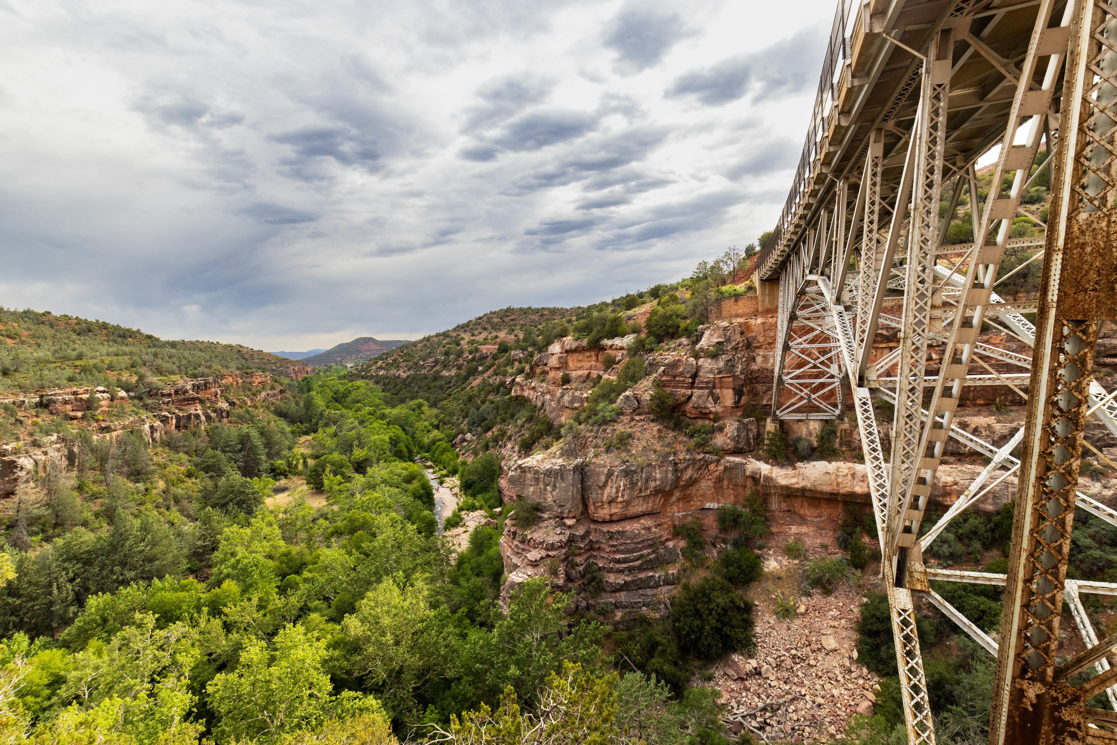 A curved metal bridge over a rocky gorge with a stream and trees underneath.