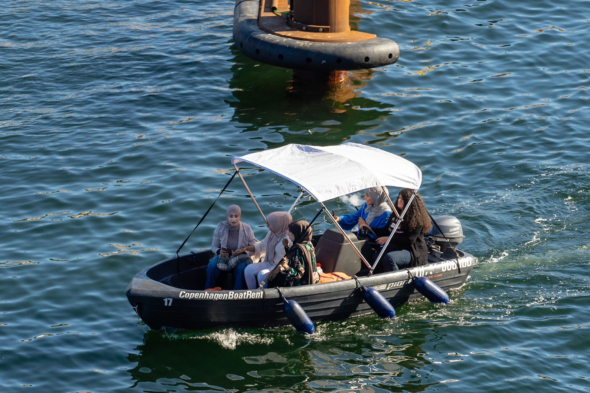 Young women wearing hijab in a boat in Copenhagen harbour, checking mobile phones and smoking water pipe