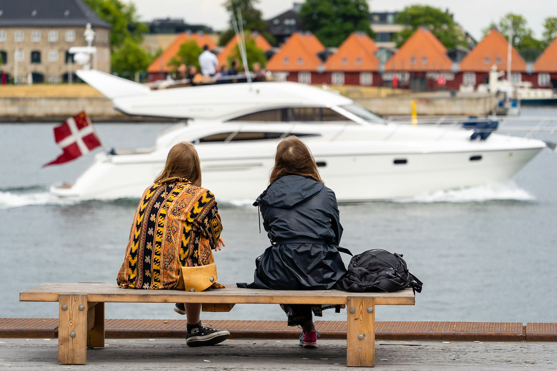 Two young women talking on bench on Ofelia Square (Ofelias Plads) by the Copenhagen waterfront, motorboat passing by