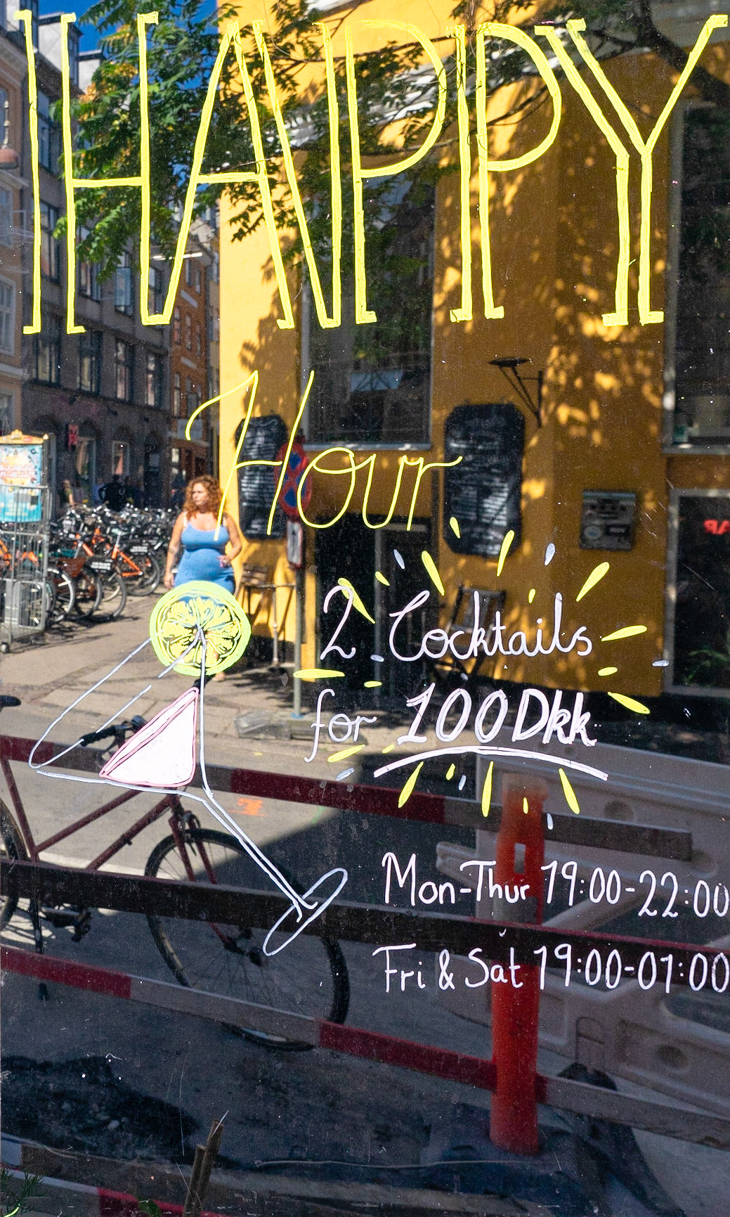 Glass sign outside hipster cafe The Living Room, Latin Quarter, Copenhagen, promoting happy hour and cocktails. Background reflected in the glass.