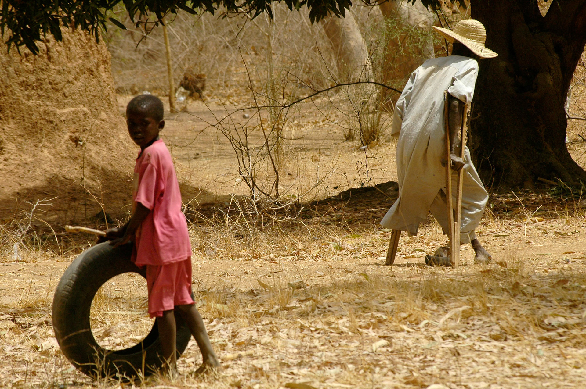 Het dagelijks leven gaat voor dit, met autoband  spelend, kind gewoon door. In de achtergrond strompelt een lepra patient op krukken. NTBLTC Lepra en TBC hospital, Zaria (Nigeria) 11 april 2006