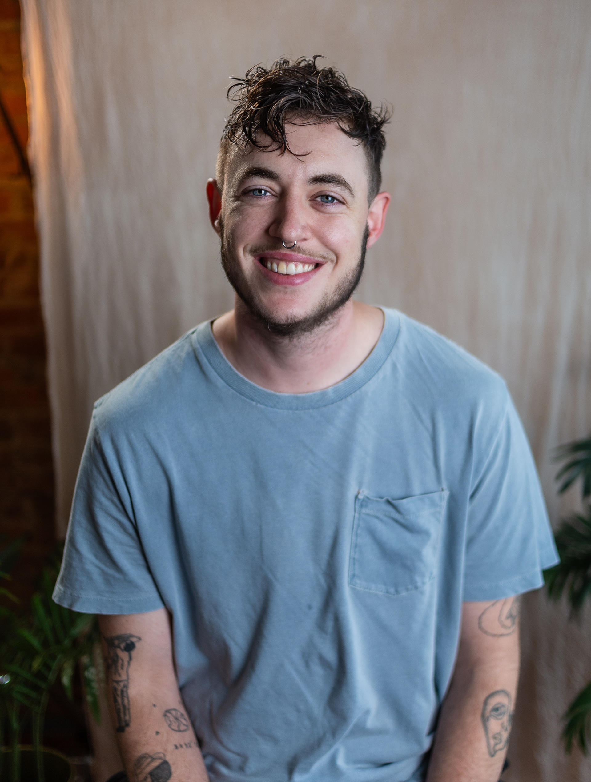 Pictured is a headshot of Mad. He's white with short brown hair and some facial hair. They're wearing a faded blue t-shirt and sitting in front of a simple tan backdrop. 