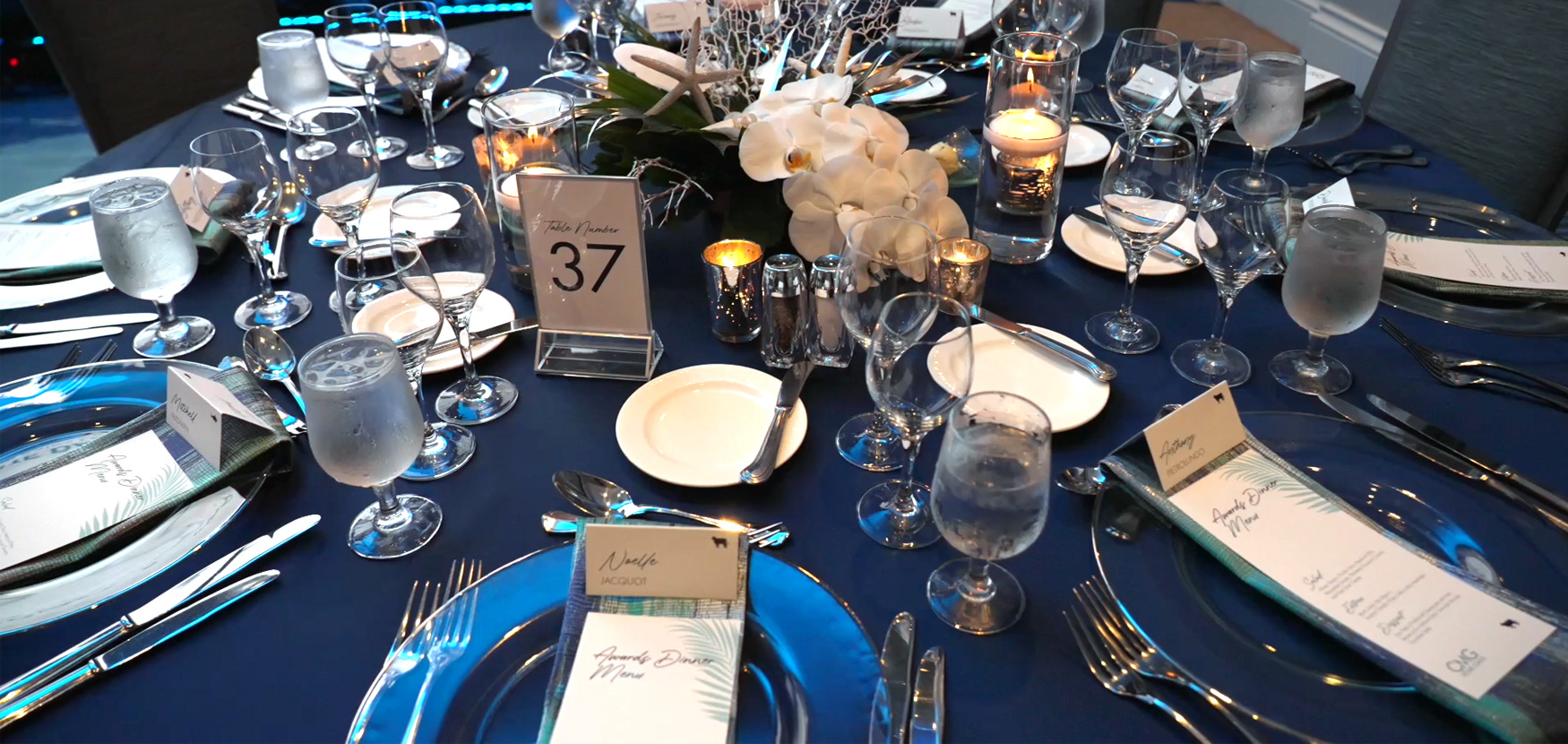 Full table setting with menus, place cards, table numbers, white orchids, candles, and dinnerwear.
