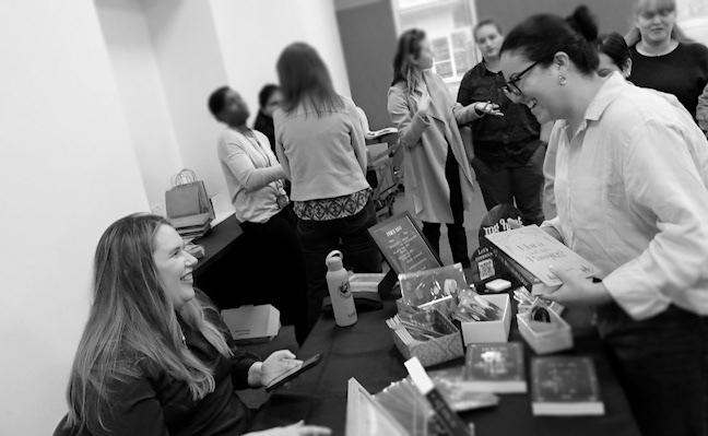 Black and white photograph of MG Hunt sitting at a table signing a copy of her book Still Waters Run Deep for a customer.