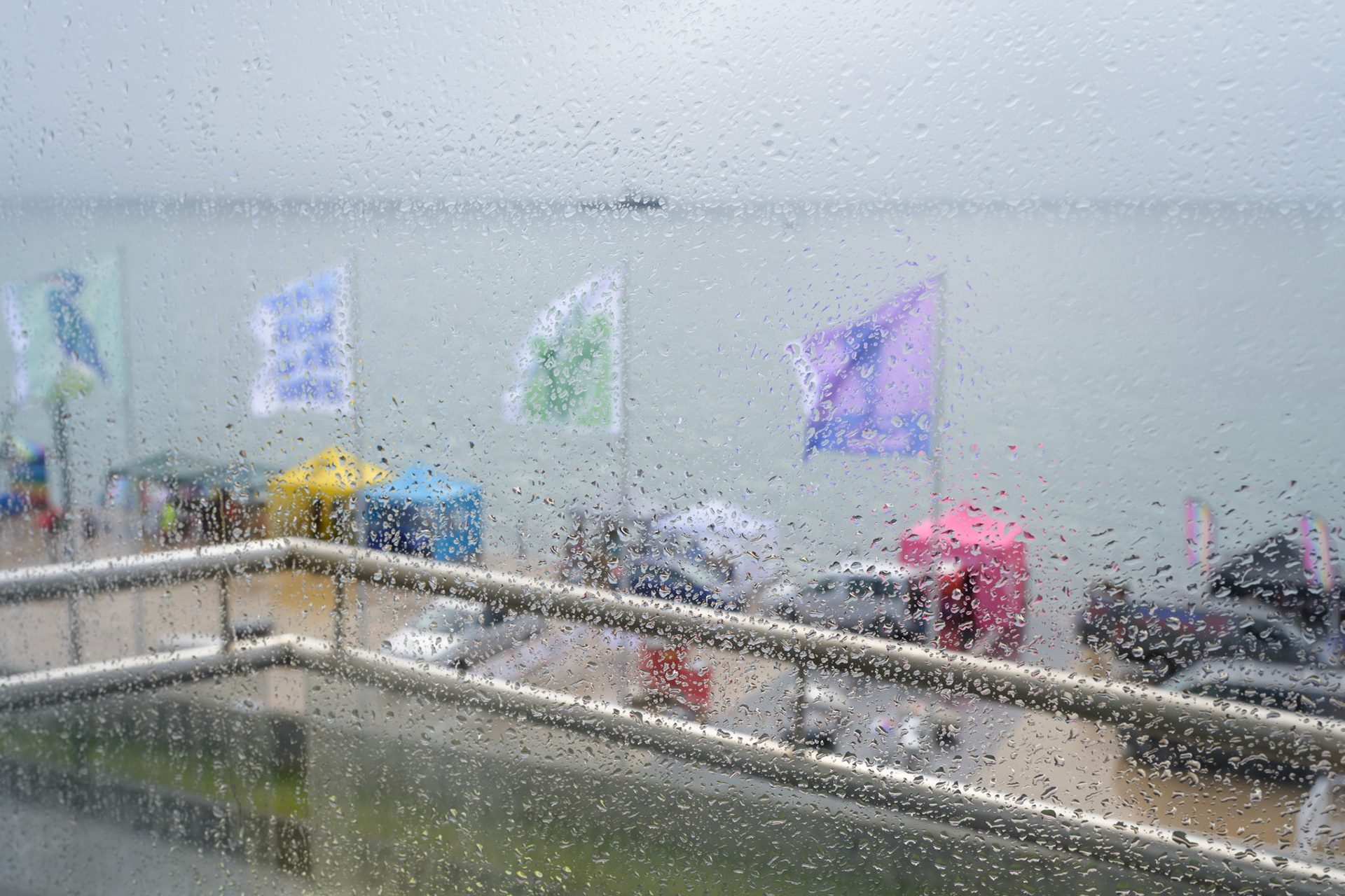 Colorful flags and booths on the sea front of Penzance during Penzance Pride festival through a window with raindrops, Cornwall