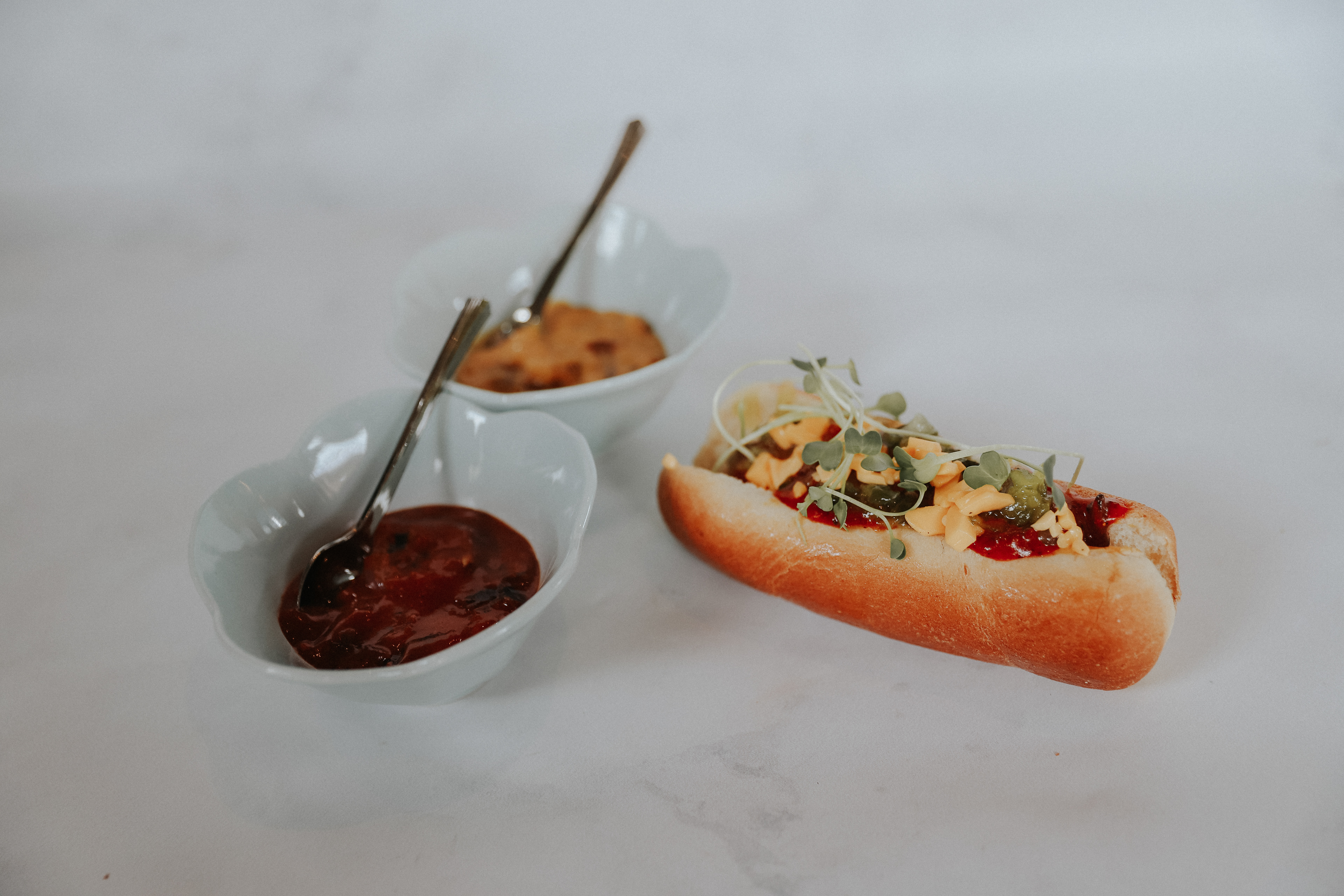 Mini Perro Americano: Salchicha, tocineta, queso cheddar rallado, pepinillos, mostaza de cebolla caramelizada, salsa de tomate con curry y brotes