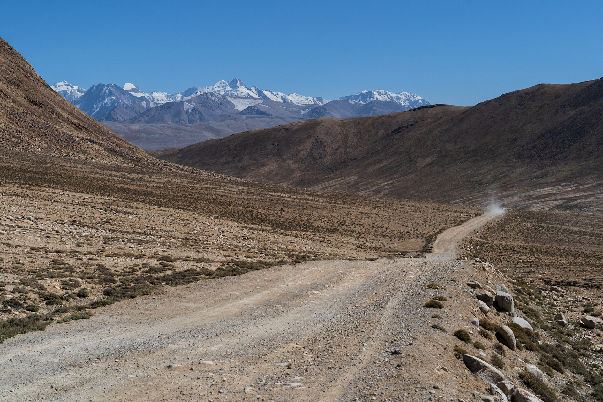 A rugged road through a barren landscape leading towards the Wakhan Valley with snowcapped peaks of Afghanistan in the background.