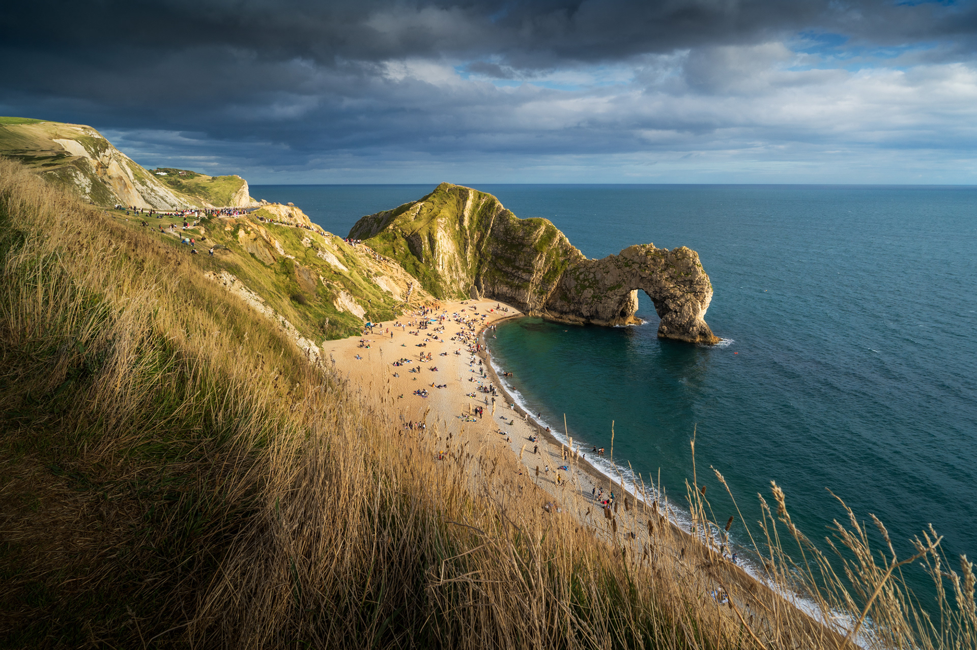 A classic view of Durde Door in Jurassic Coast, Dorset, England, with grasses in the foreground and a dramatic sky with golden light hitting the landscape.
