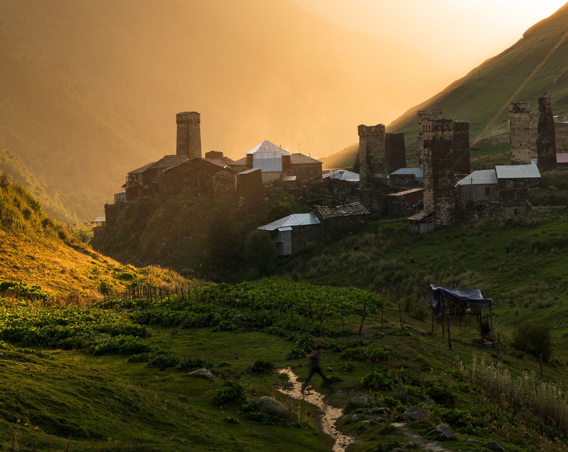 A bright orange sunset in a mountain village, with a farm in the foreground and defensive stone towers in the background.