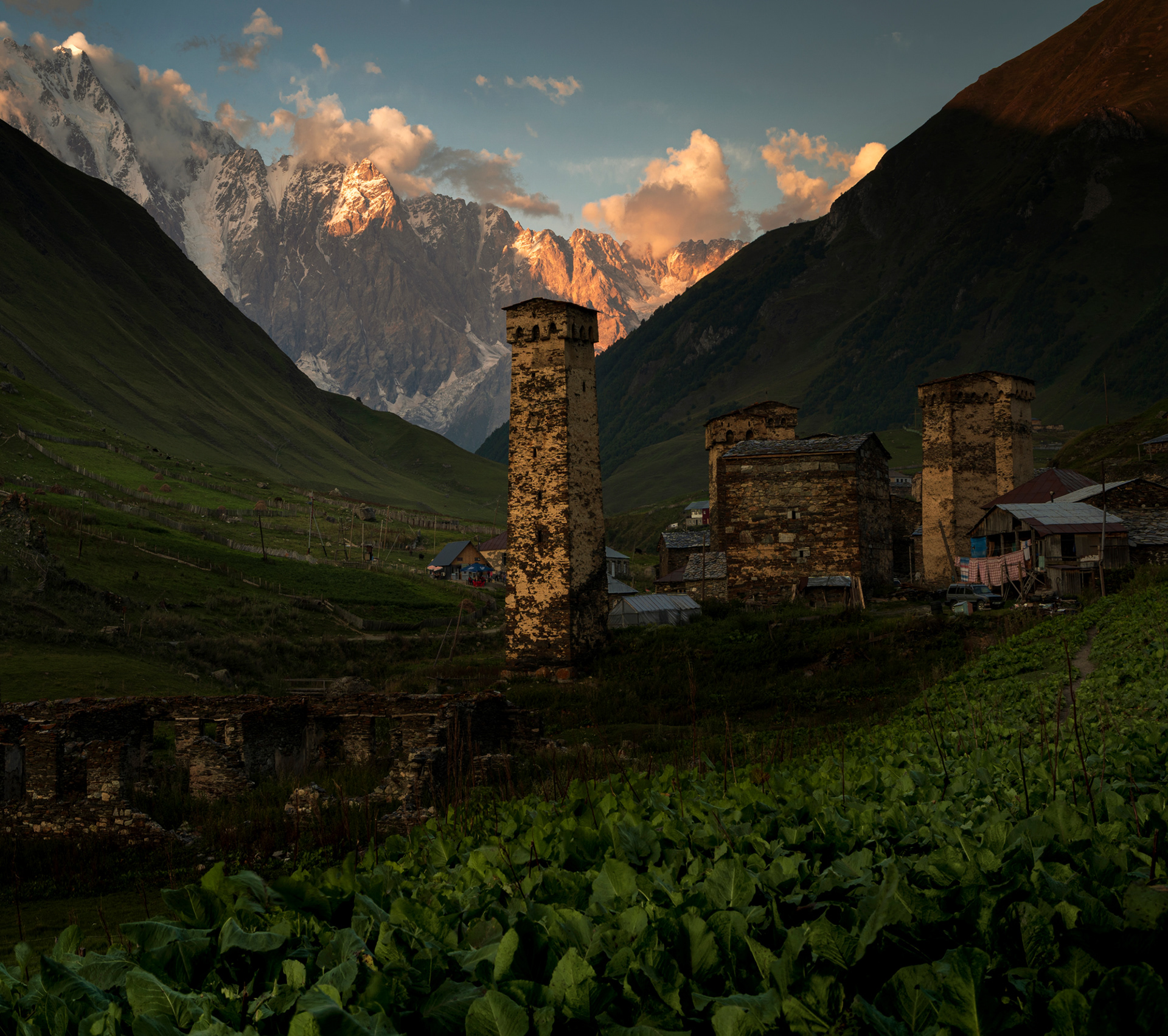 A bright orange sunset in a mountain village, with a farm in the foreground and defensive stone towers in the background.