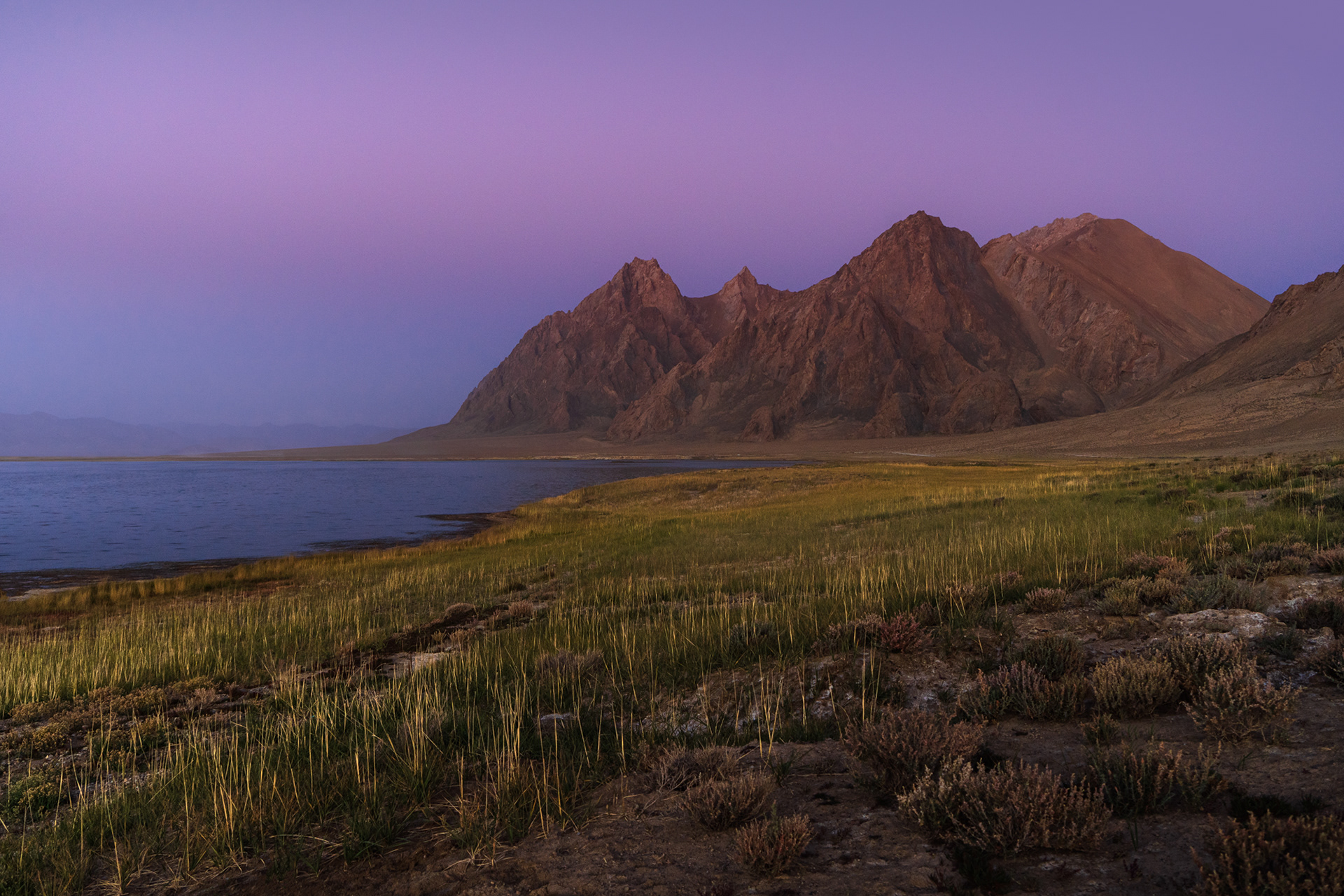Dusk above a mountainous terrain above a lake and grassy shores.