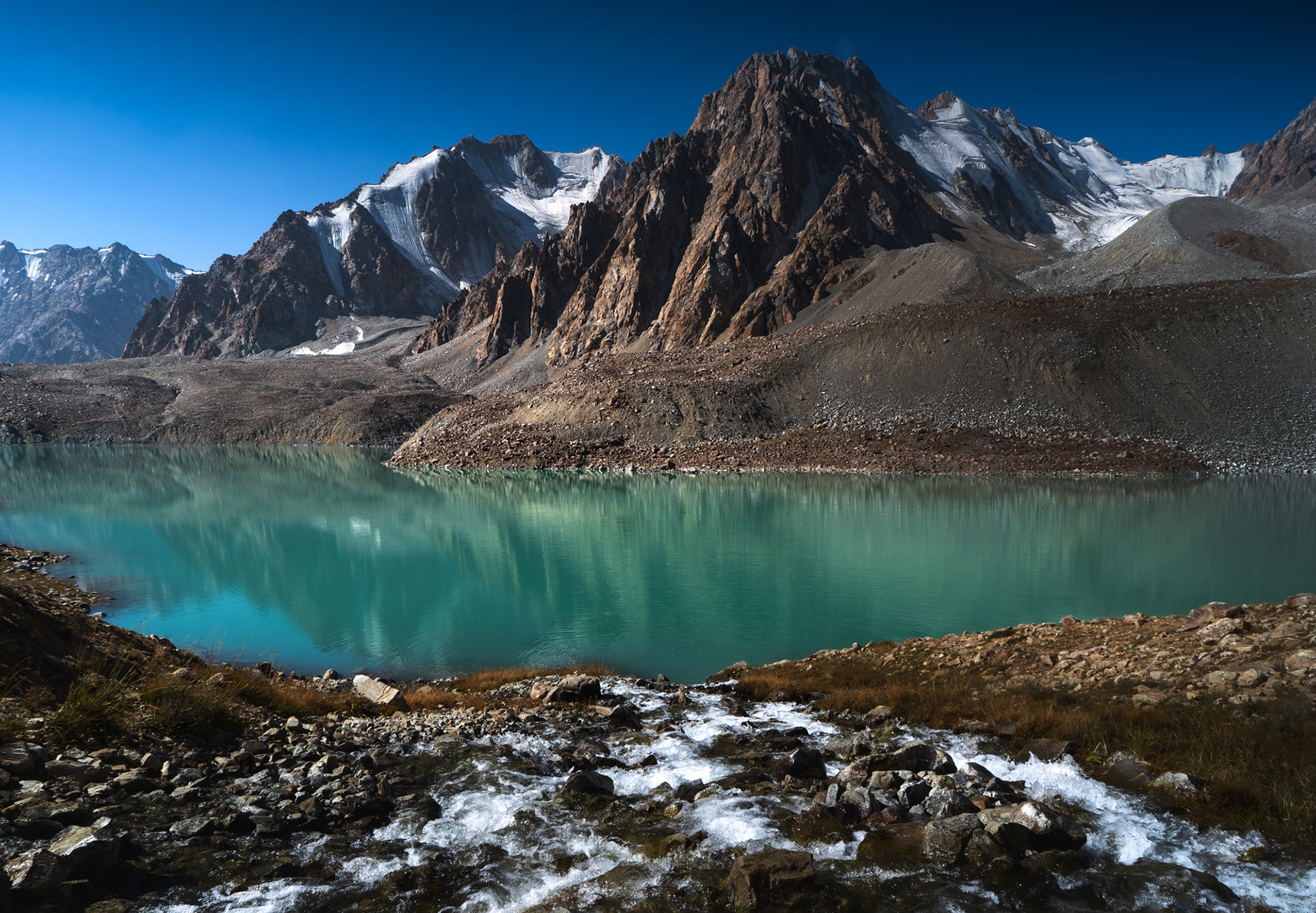 A mountain stream flows into the emerald Tsaxinkul Lake with epic peaks rising above it.