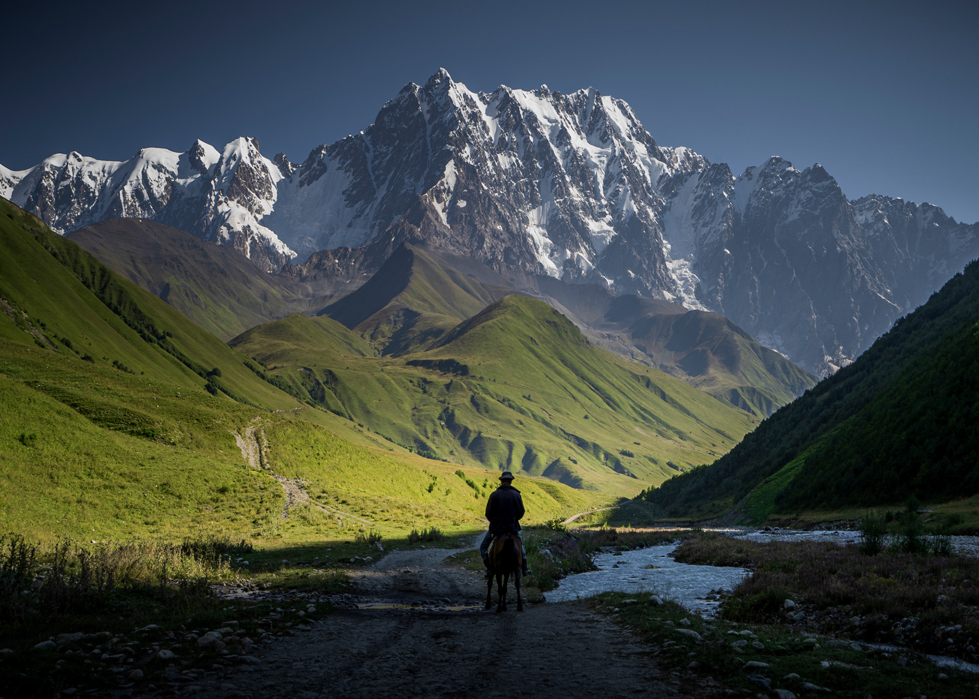 A photo of a man on a horse in front of a large, lush green mountain with snow on top.