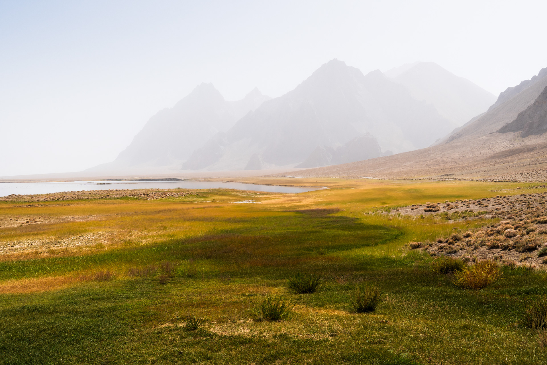 A hazy day in a rugged mountainous terrain, with colourful grasslands and a lake.