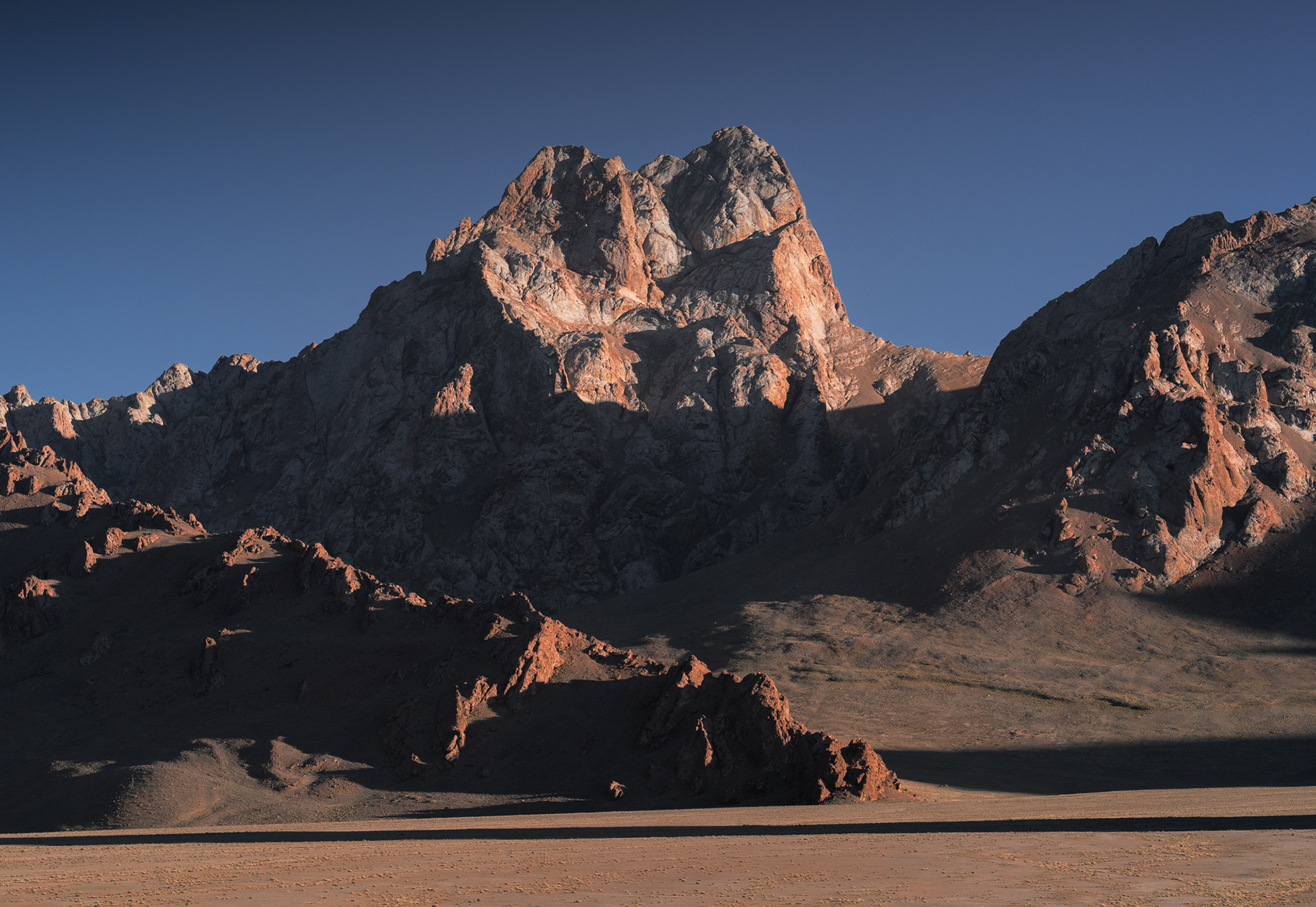 A sharp, orange mountain rises from a flat valley in Tajikistan under a clear sky.