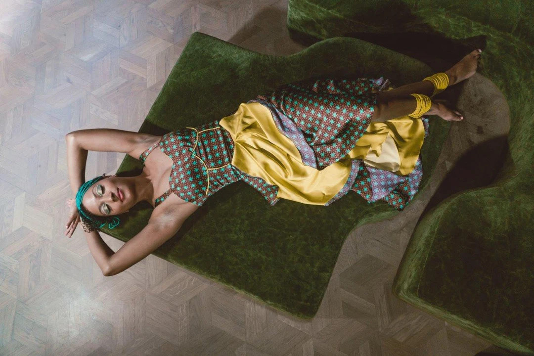 mujer recostada sobre un sillón usando un vestido estampado con diseños de inspiración africana coordinados, en colores bordeaux, amarillo y turquesa