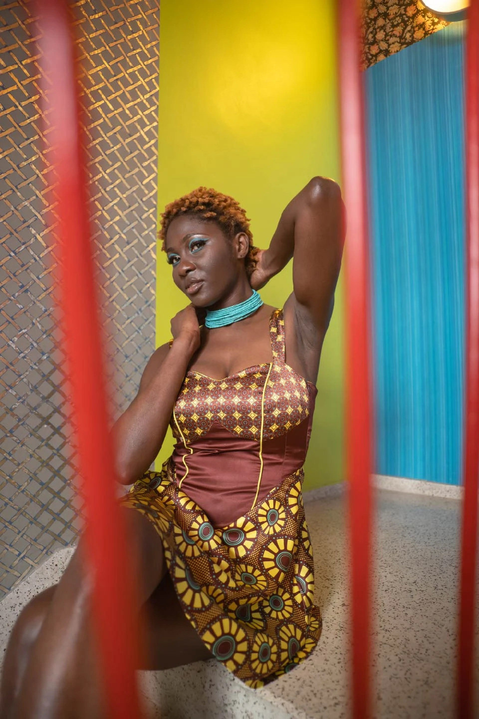 mujer sentada, mirando a la cámara, luciendo un vestido corto estampado con diseños de inspiración africana coordinados, en colores bordeaux, amarillo y turquesa
