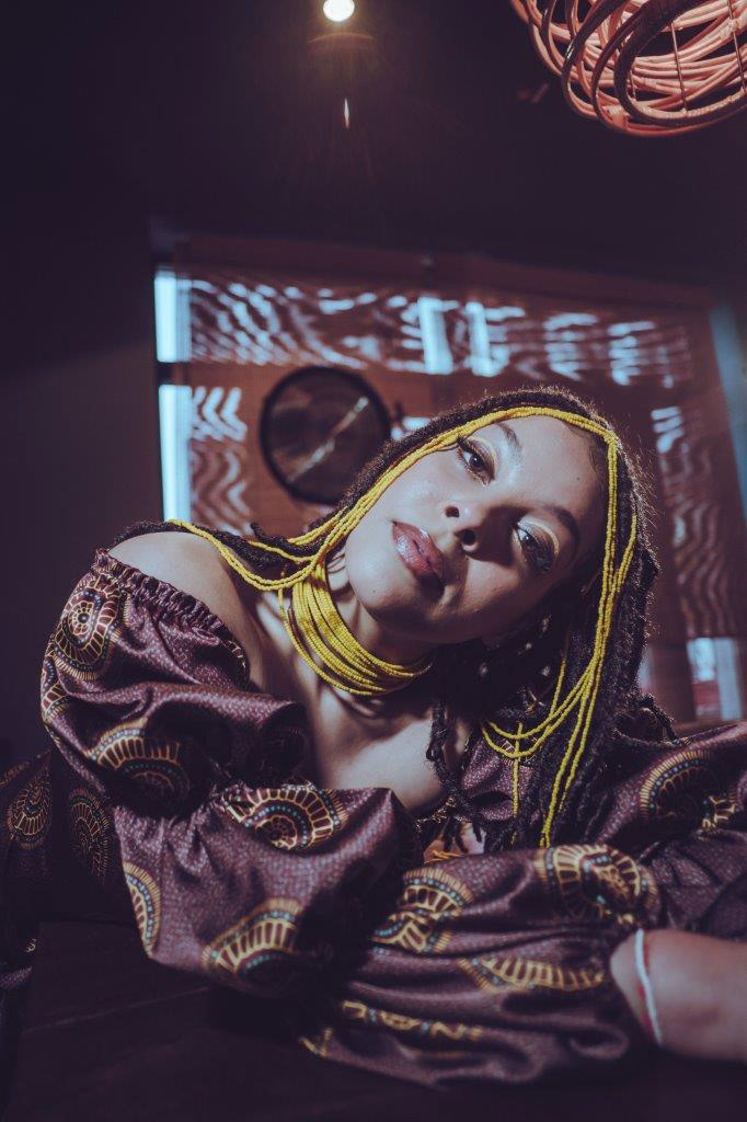 Mujer mirando directamente a la cámara, luciendo un vestido estampado con un diseño de estilo africano, en colores bordeaux, amarillo y detalles en turquesa
